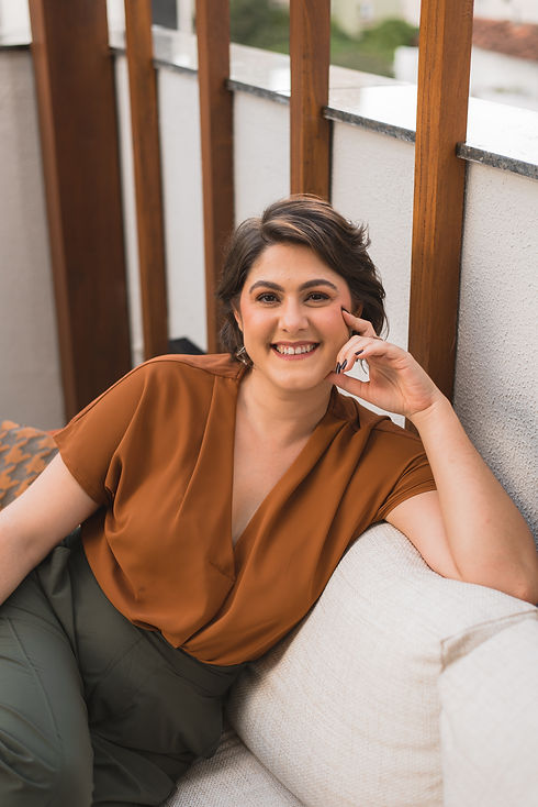 Mulher de cabelos curtos sorridente com a mão no rosto, sentada em um sofá de tecido claro. A mulher veste uma camisa marrom e uma calça verde.