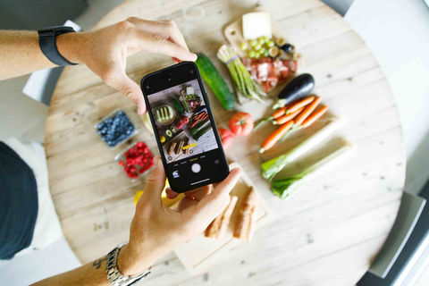 An image of someone holding a mobile phone to take a photo of fresh vegetables laid out neatly on a wooden board