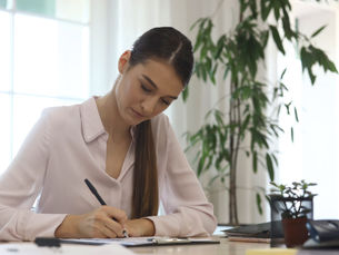 Woman in a light blouse writing at a desk with a pen. A potted plant and office supplies are on the table. Bright window in the background.