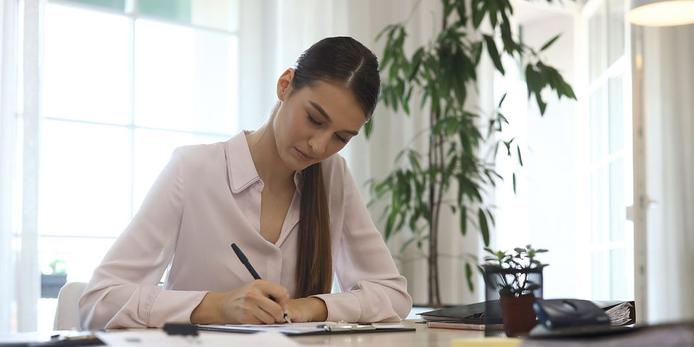Woman in a light blouse writing at a desk with a pen. A potted plant and office supplies are on the table. Bright window in the background.