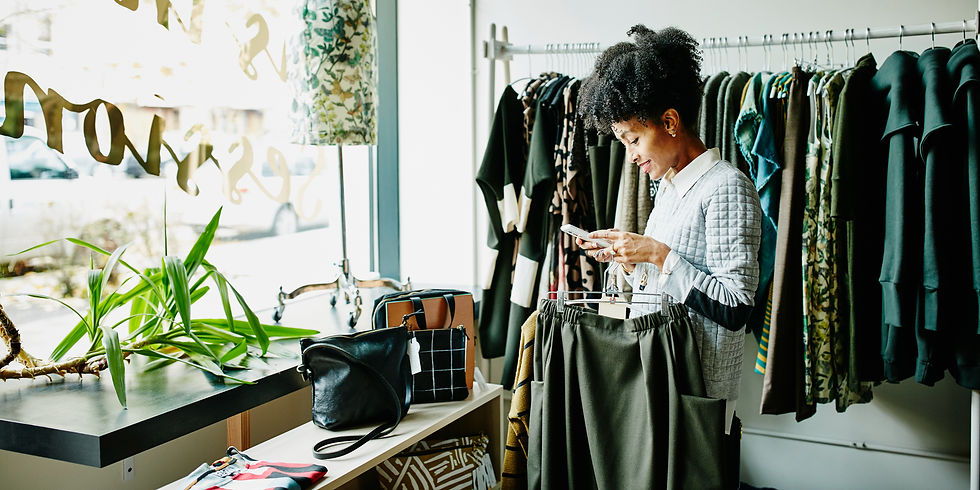 Woman business owner in a boutique on a phone, examining clothes on a rack. Bright setting with natural light, green plants, and visible handbags.