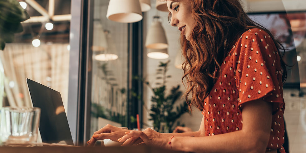 Red-haired woman in a polka dot dress types on a laptop at a café. Warm light, hanging lamps, and plants create a cozy, focused mood.