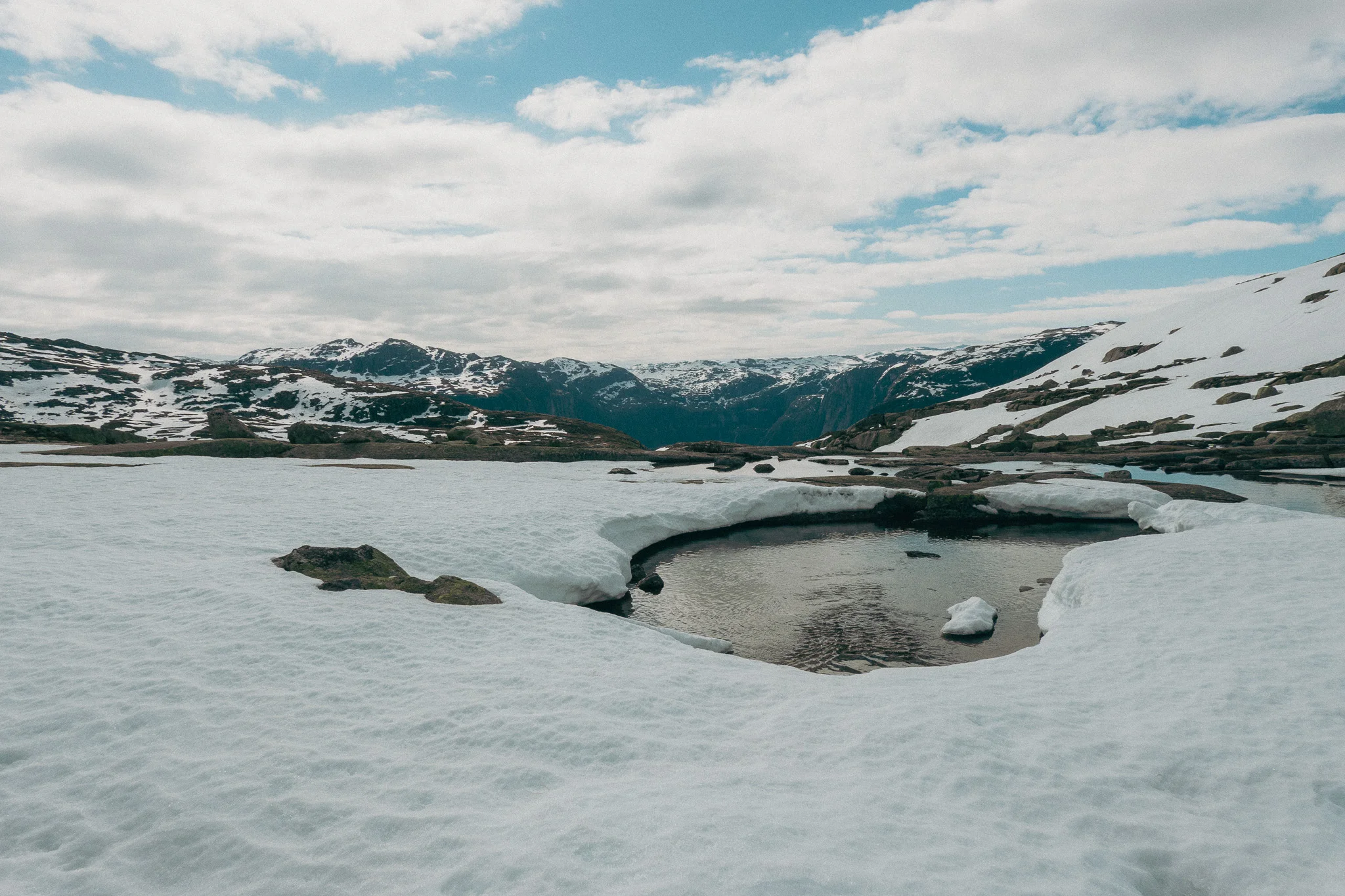 A small body of water surrounded by snow with snowcapped mountains in the distance beneath partly cloudy skies on the Trolltunga trail in Norway