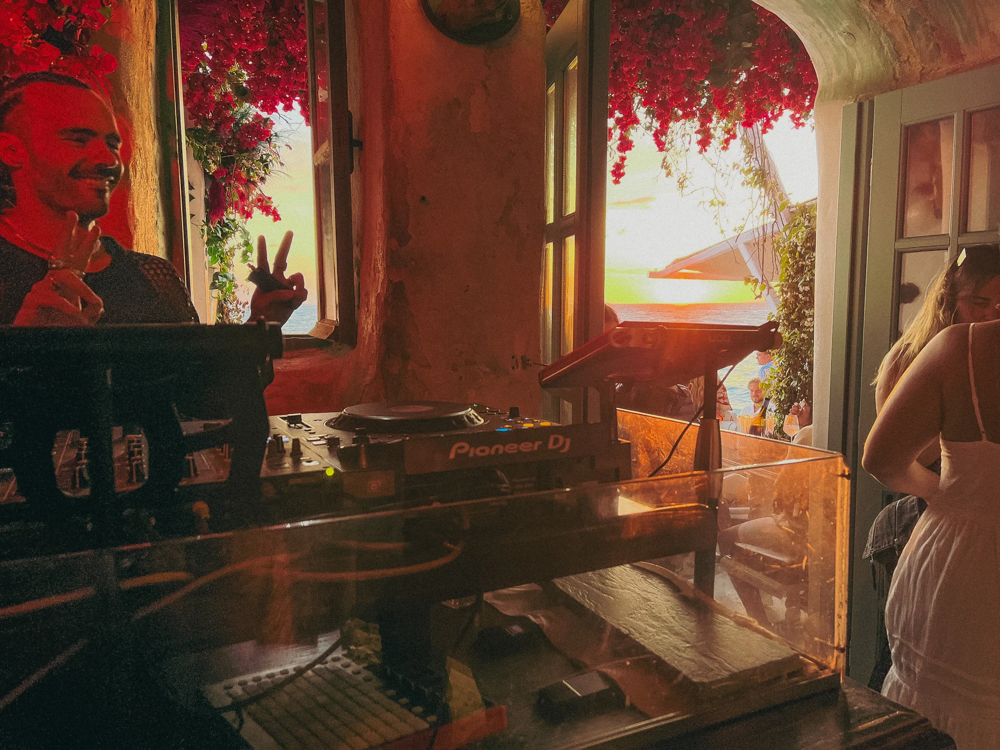 A DJ poses smiling and flashing the peace sign surrounded by red roses a decoration and a window filled with sunlight