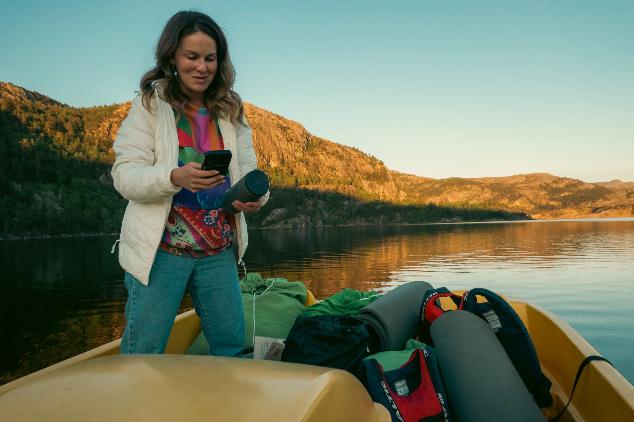 A woman holding a phone and a speaker standing in a paddle boat full of gear on a lake with mountains in the background at sunset.