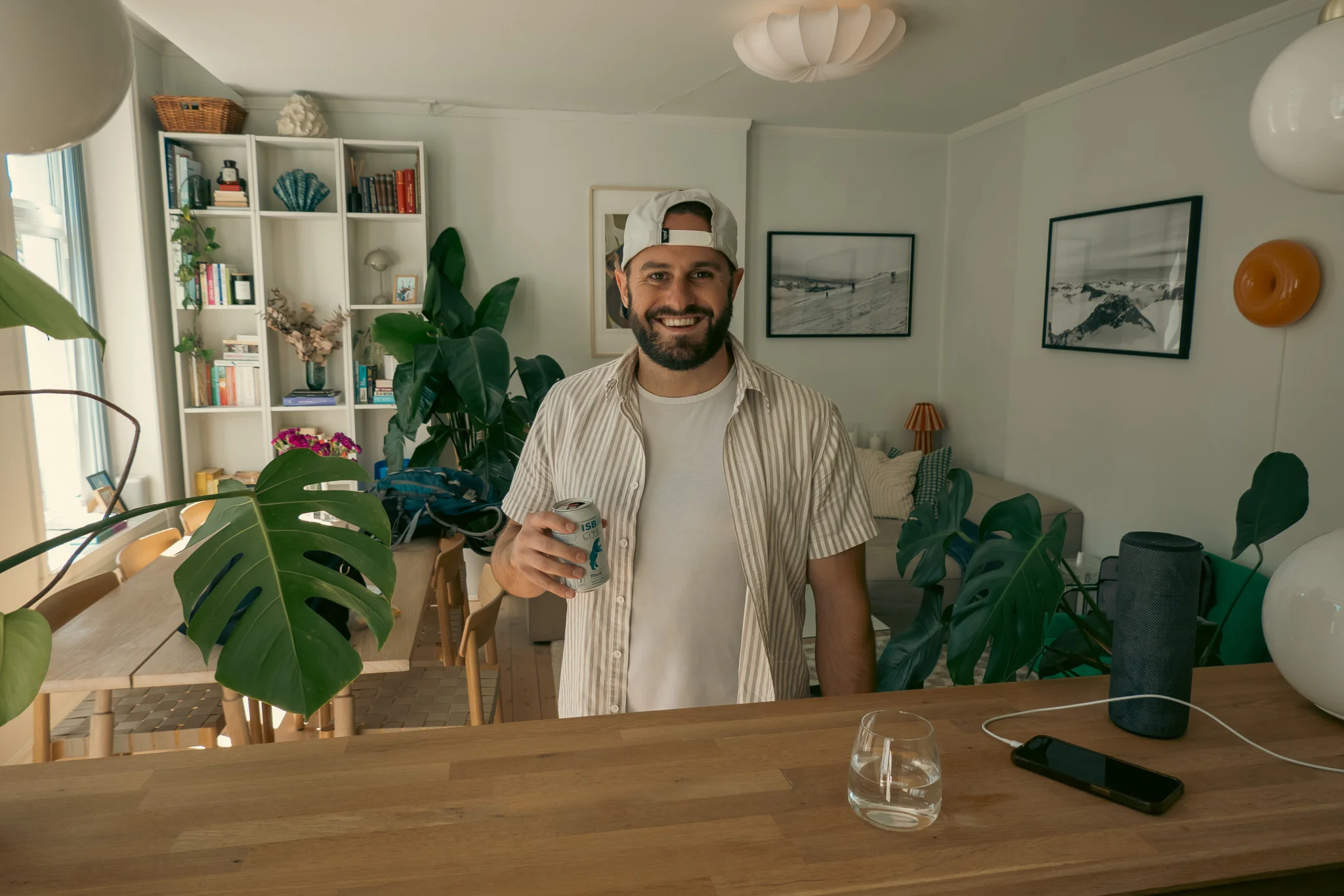 Doug holding a beer in an apartment in Oslo, Norway surrounded by green plants and a counter in the foreground with a glass of water, iPhone charging and Bluetooth speaker on it. 