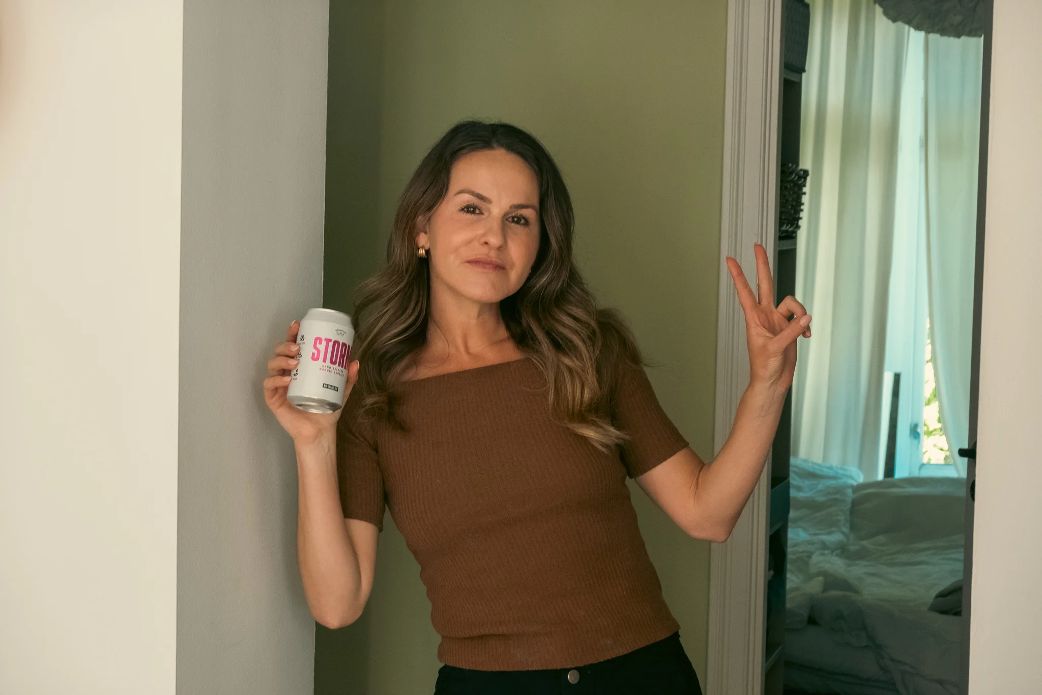 Kelly in a hallway in an apartment in Oslo, Norway holding a beer in one hand and holding up the peace sign with the other.