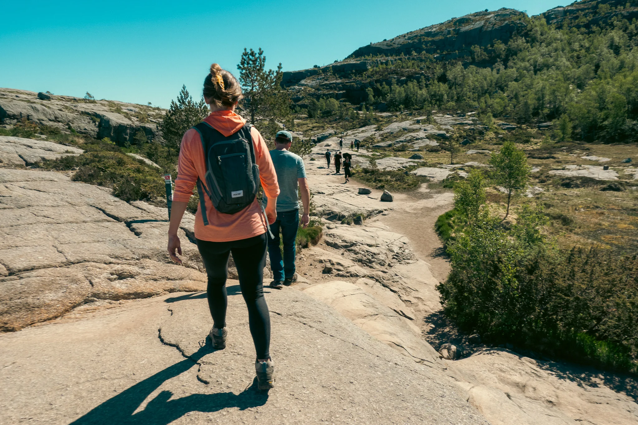 A man and woman walk along a rocky hiking trail with trees and mountains in the background giving a sense of adventure.