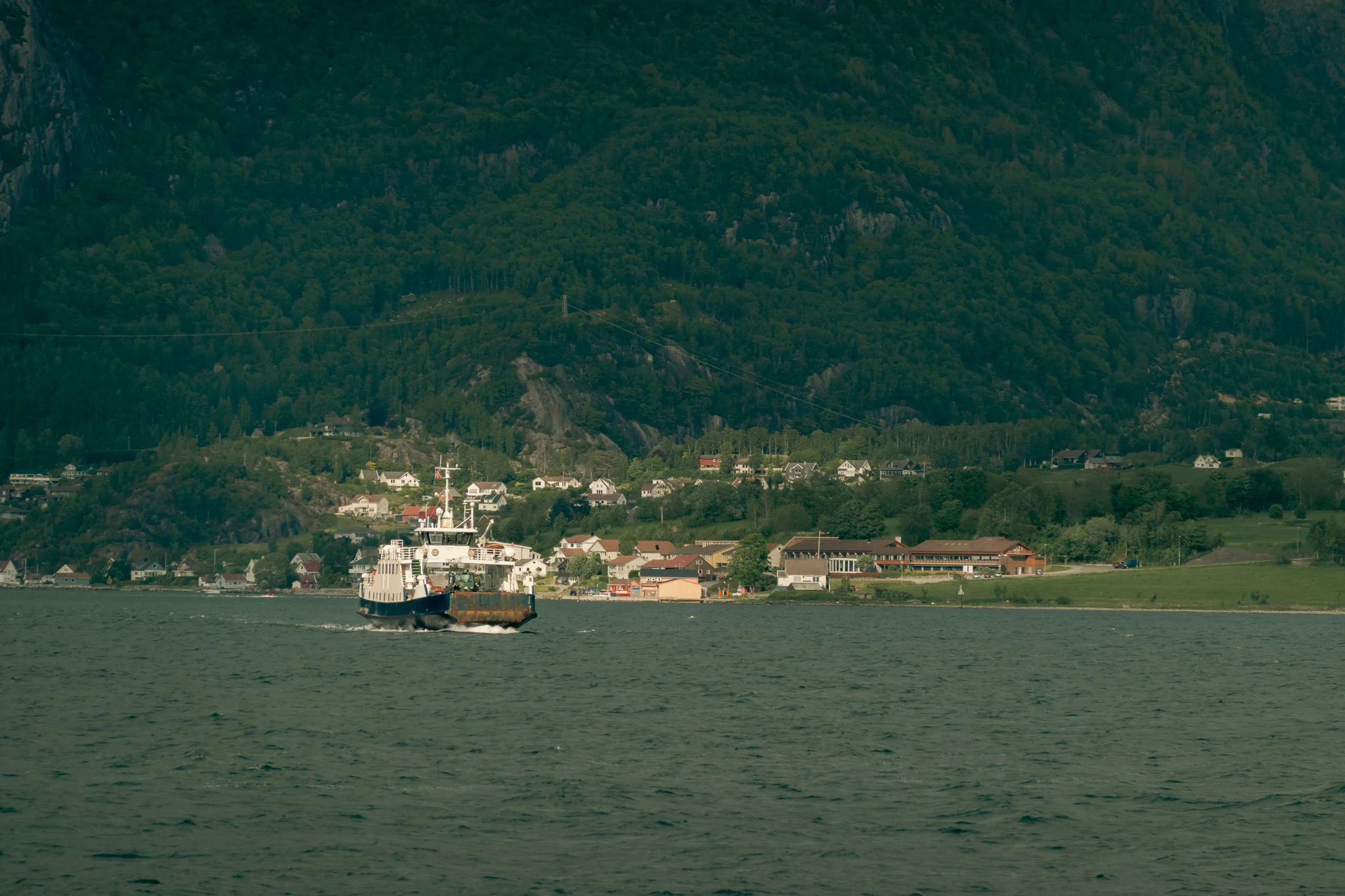 A ferry motoring along a fjord with trees and mountains in the background.
