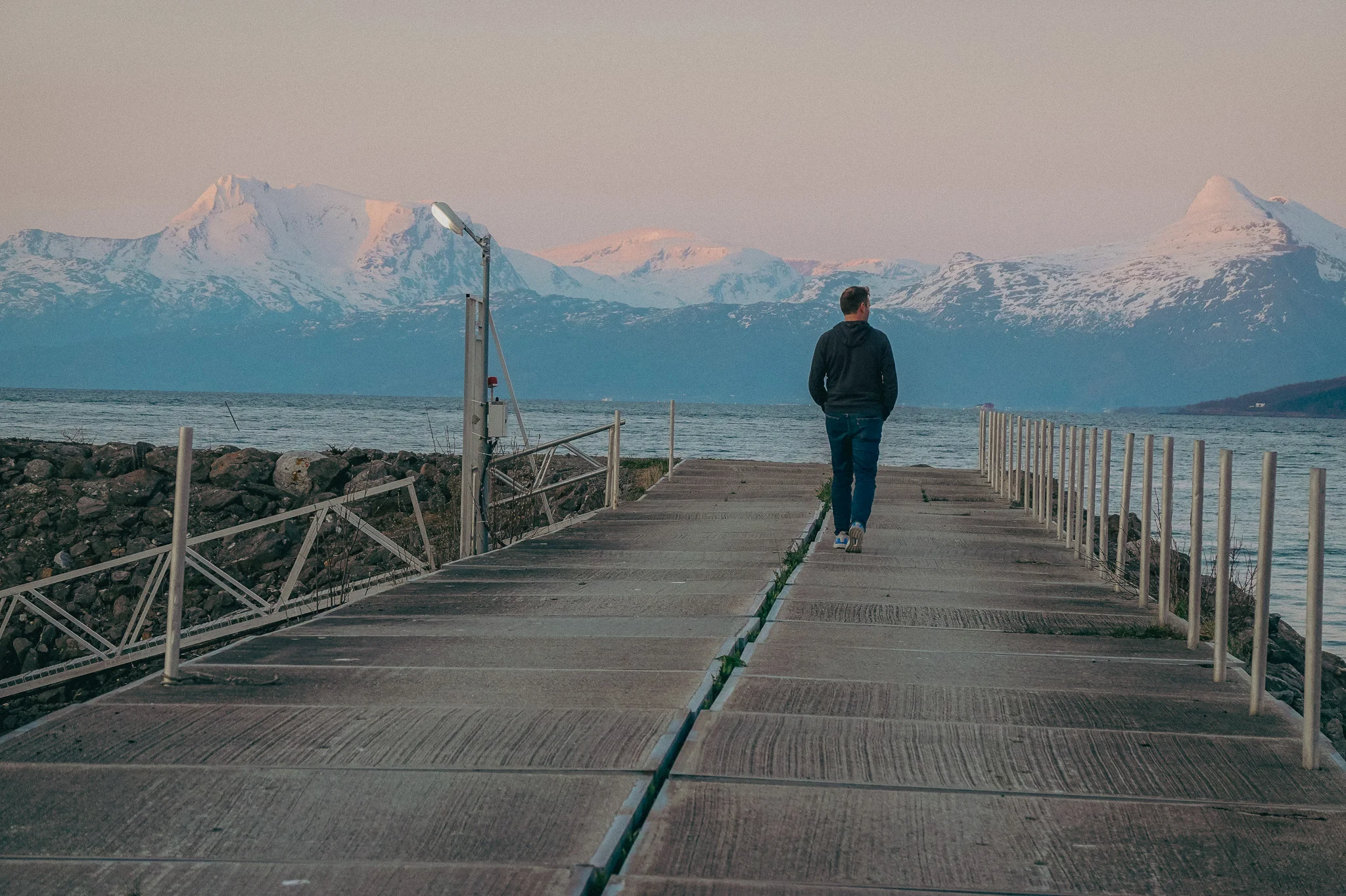 A man strolls down a pier looking off into the distance with snow-capped mountains and water in the background
