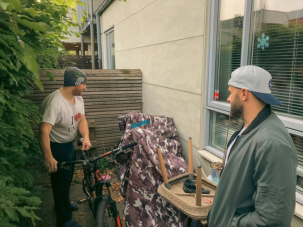 Donat and Doug outside beside a covered bike. One holds a basket with legs and cable. Green foliage and a window with a snowflake sticker in view.