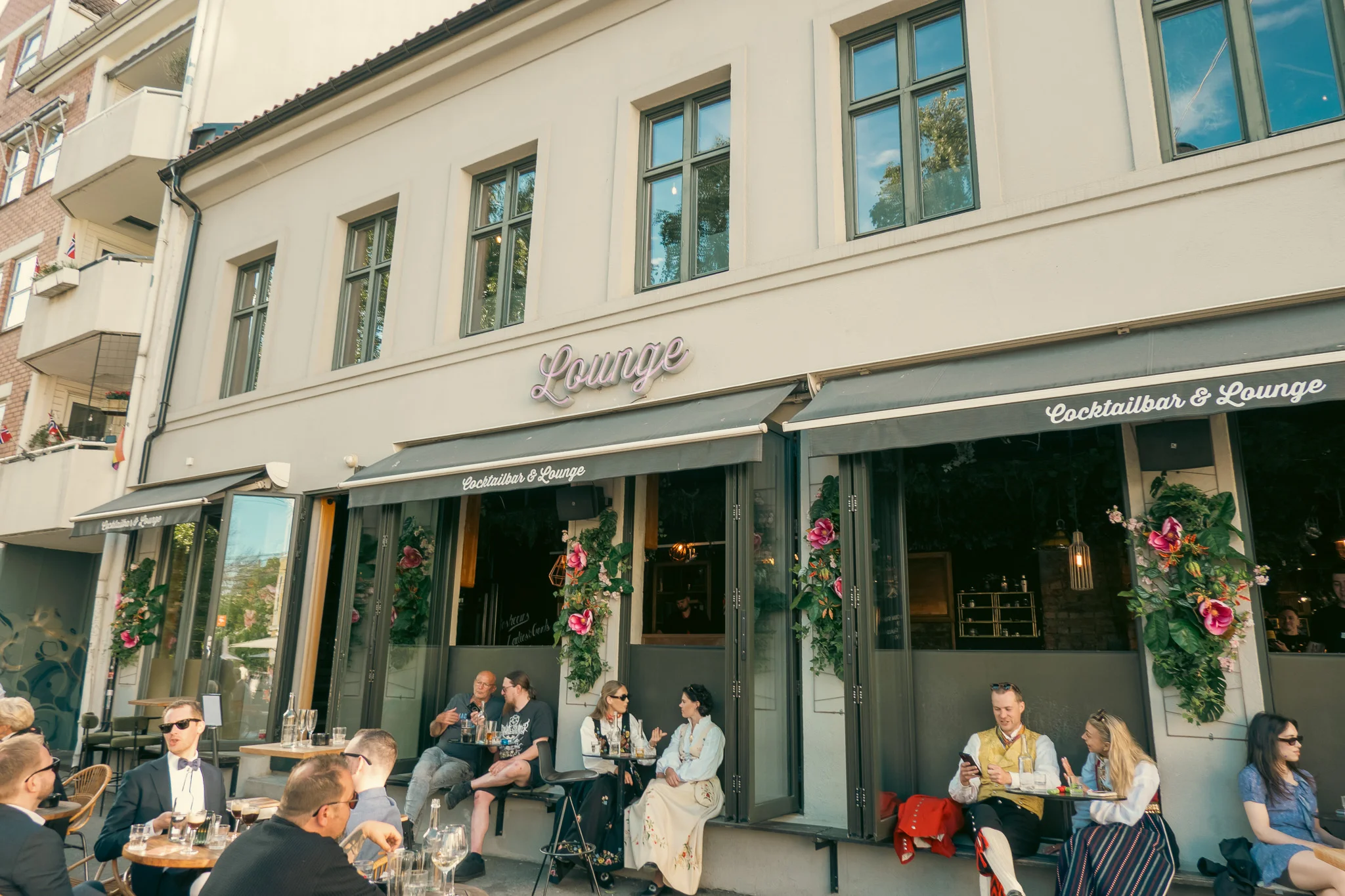 Several groups of people sitting outside a restaurant on the sidewalk enjoying drinks and celebrating Constitution Day in Oslo Norway