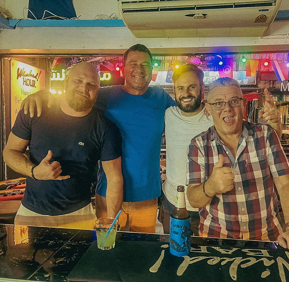 Four smiling men pose at a bar with colorful lights. Two drinks are on the counter, and a "Wicked Hour!" sign is visible in the background.