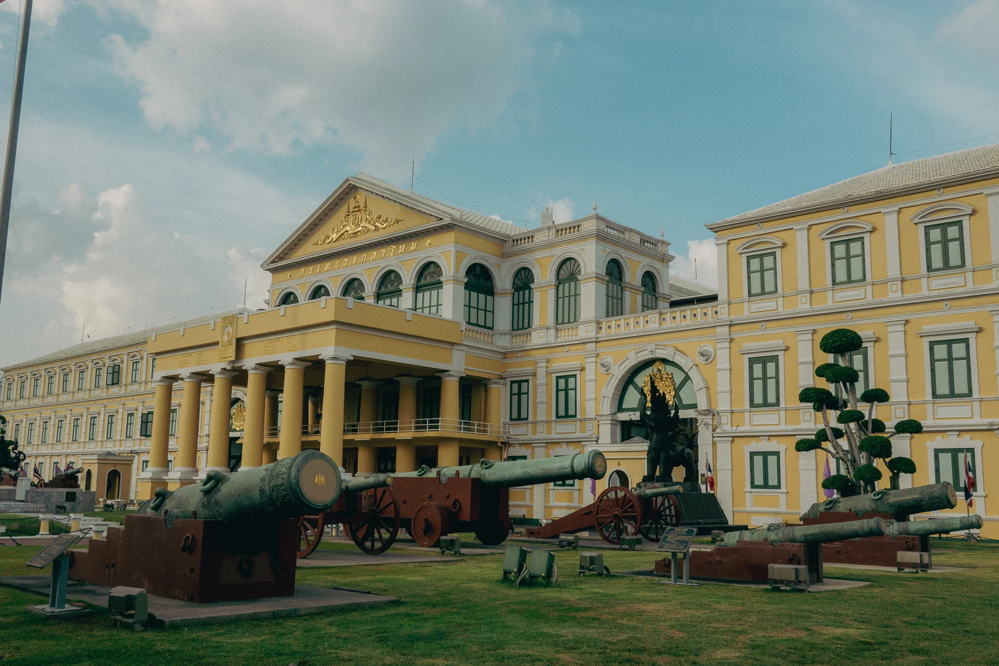 Large yellow state building in Bangkok with green grass and cannons in the front and blue skies in the background