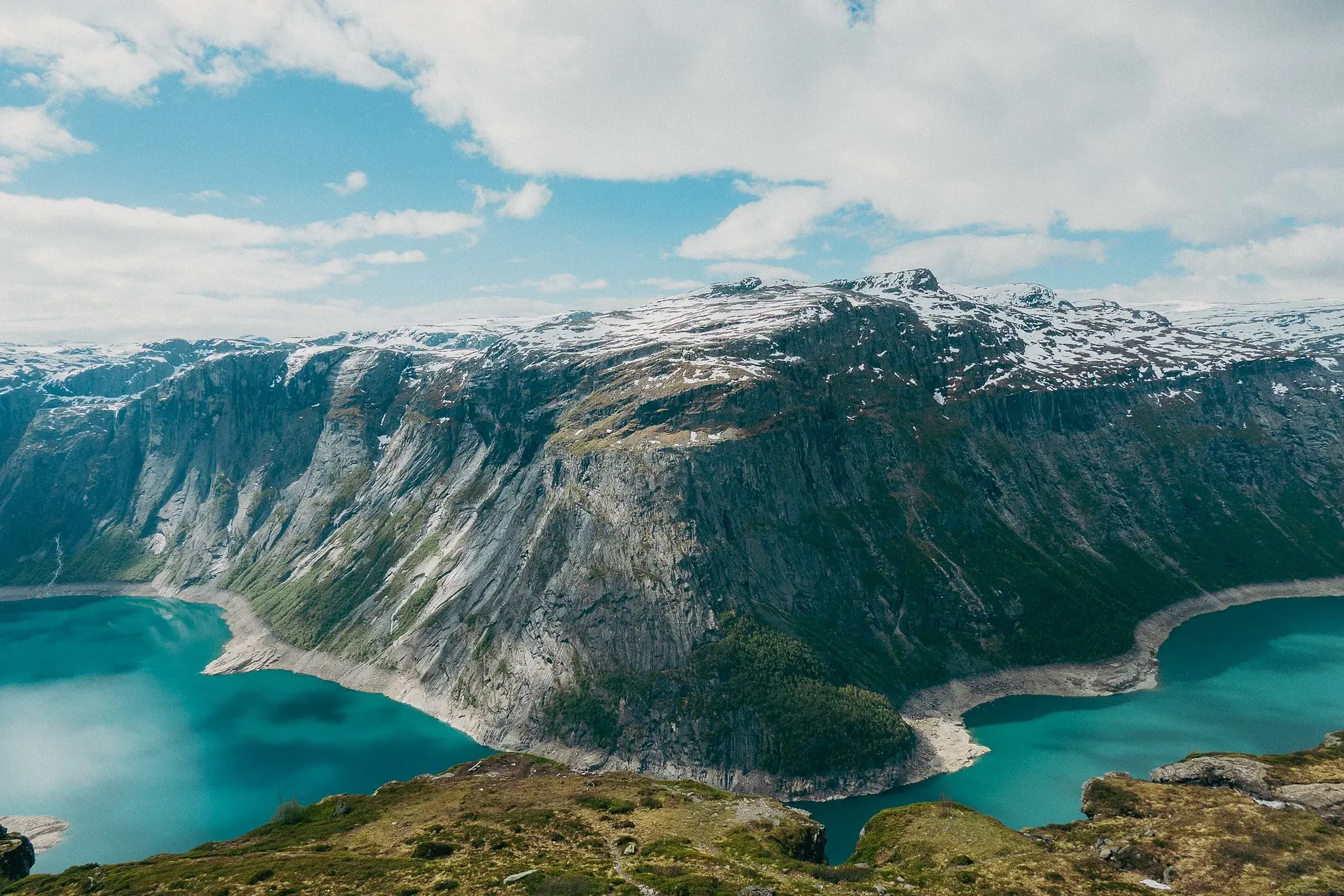Scenic landscape with rocks and grass in the foreground and a blue lake and snowcapped mountains beneath a blue sky on the Trolltunga trail in Norway