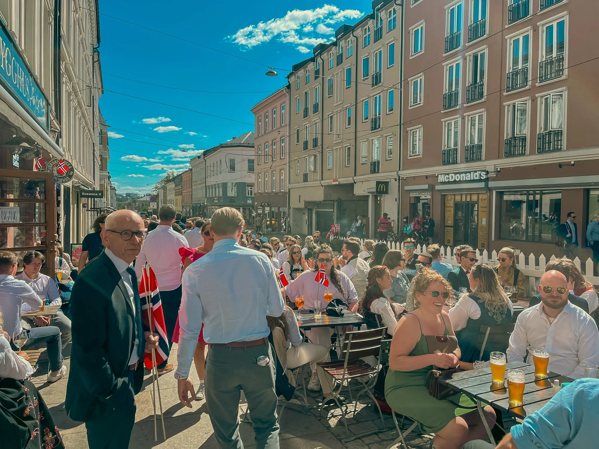 A large group of people celebrating Constitution Day at tables outside on the sidewalk in Oslo, Norway.