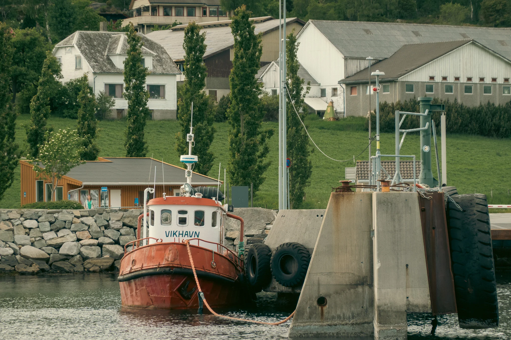A red tug boat docked at a cement pier with green trees and a white building in the background.