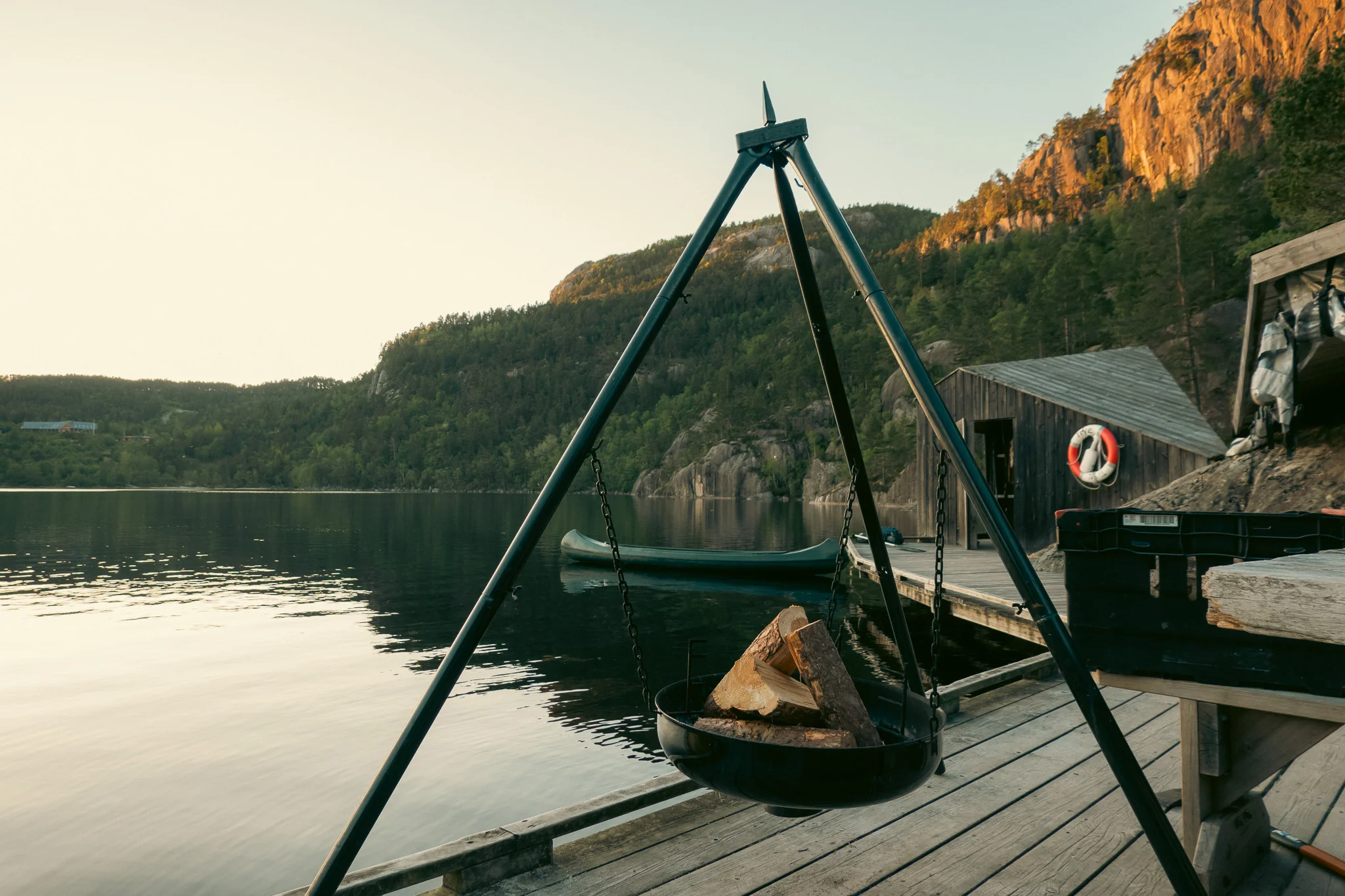 A fire pit filled with wood on a dock alongside a like with a canoe, small hut, and tree-lined mountains in the background.
