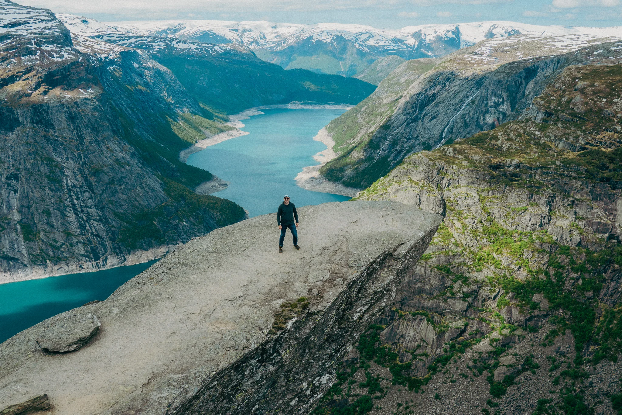 Adult male stands at the tip of Trolltunga in Norway with snowcapped mountains and blue water in the background