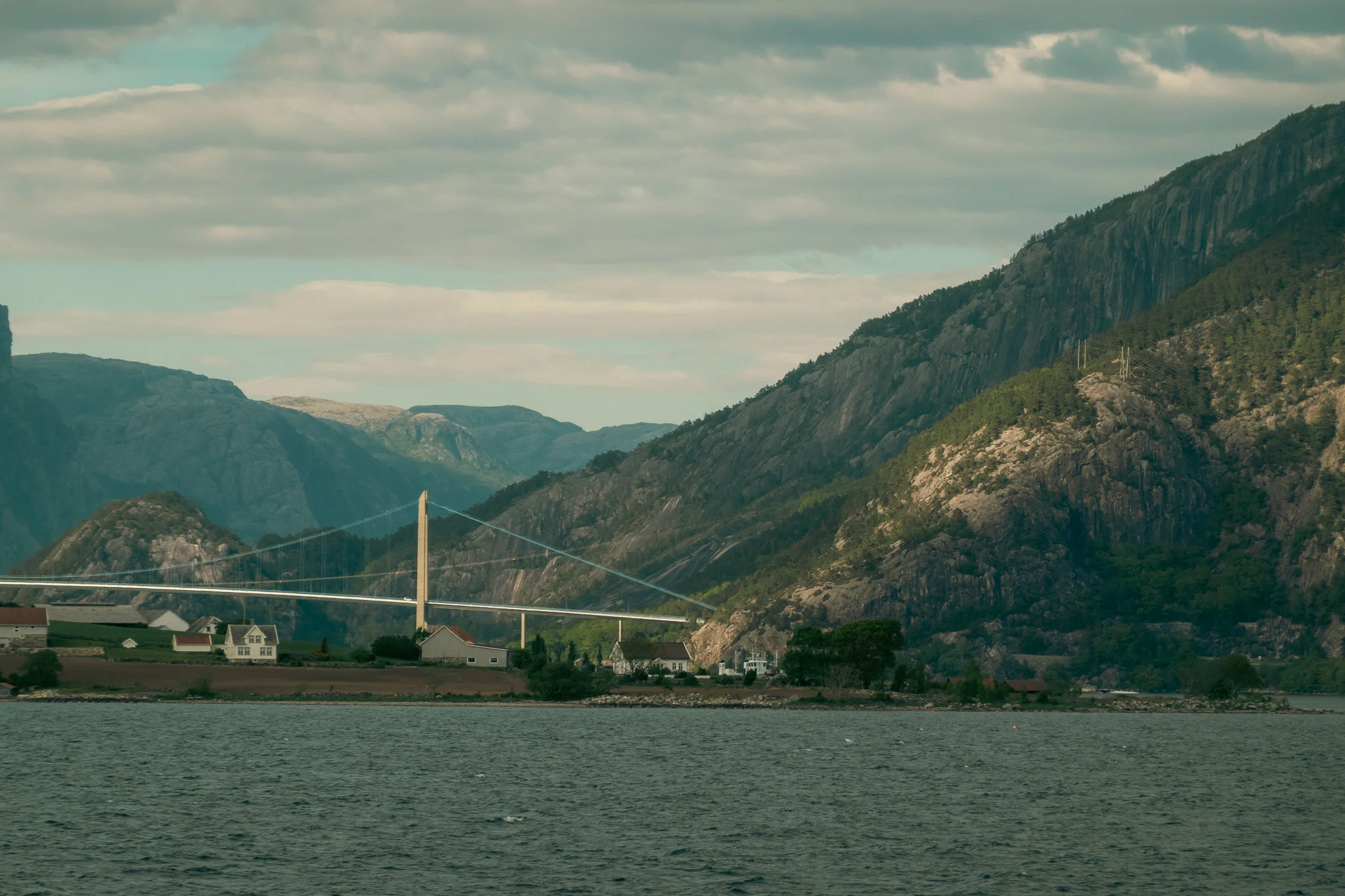A bridge crossing a fjord with layers of mountains in the background on a cloudy day.