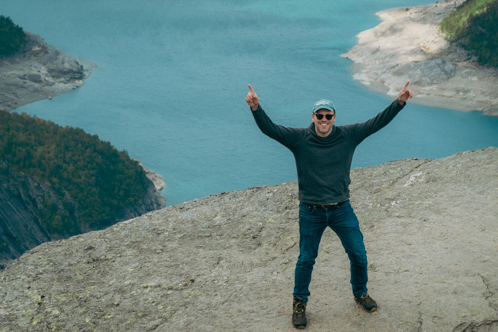 Adult male stands at the tip of Trolltunga in Norway with snowcapped mountains and blue water in the background