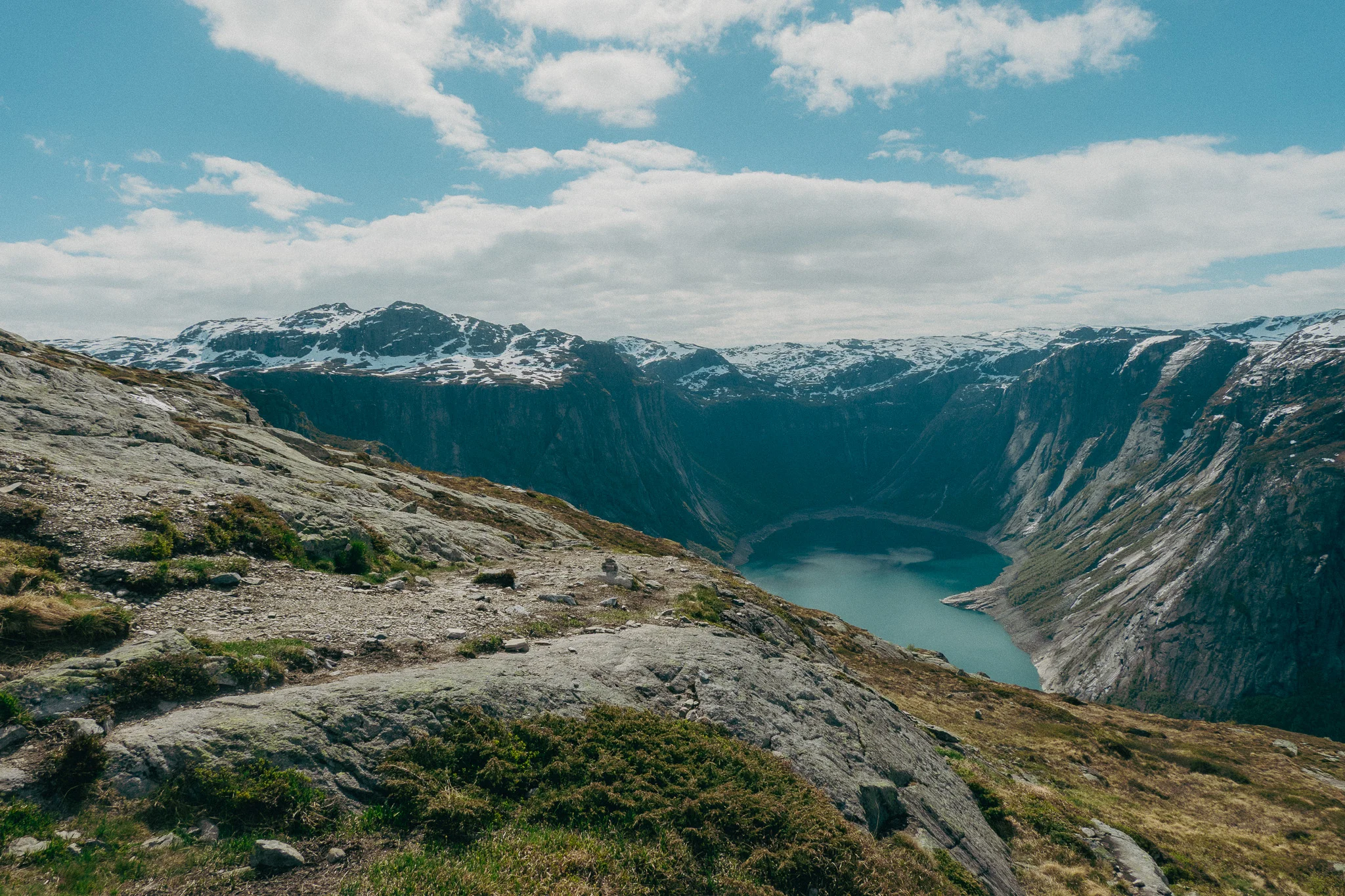 Scenic landscape with rocks and grass in the foreground and a blue lake and snowcapped mountains beneath a blue sky on the Trolltunga trail in Norway