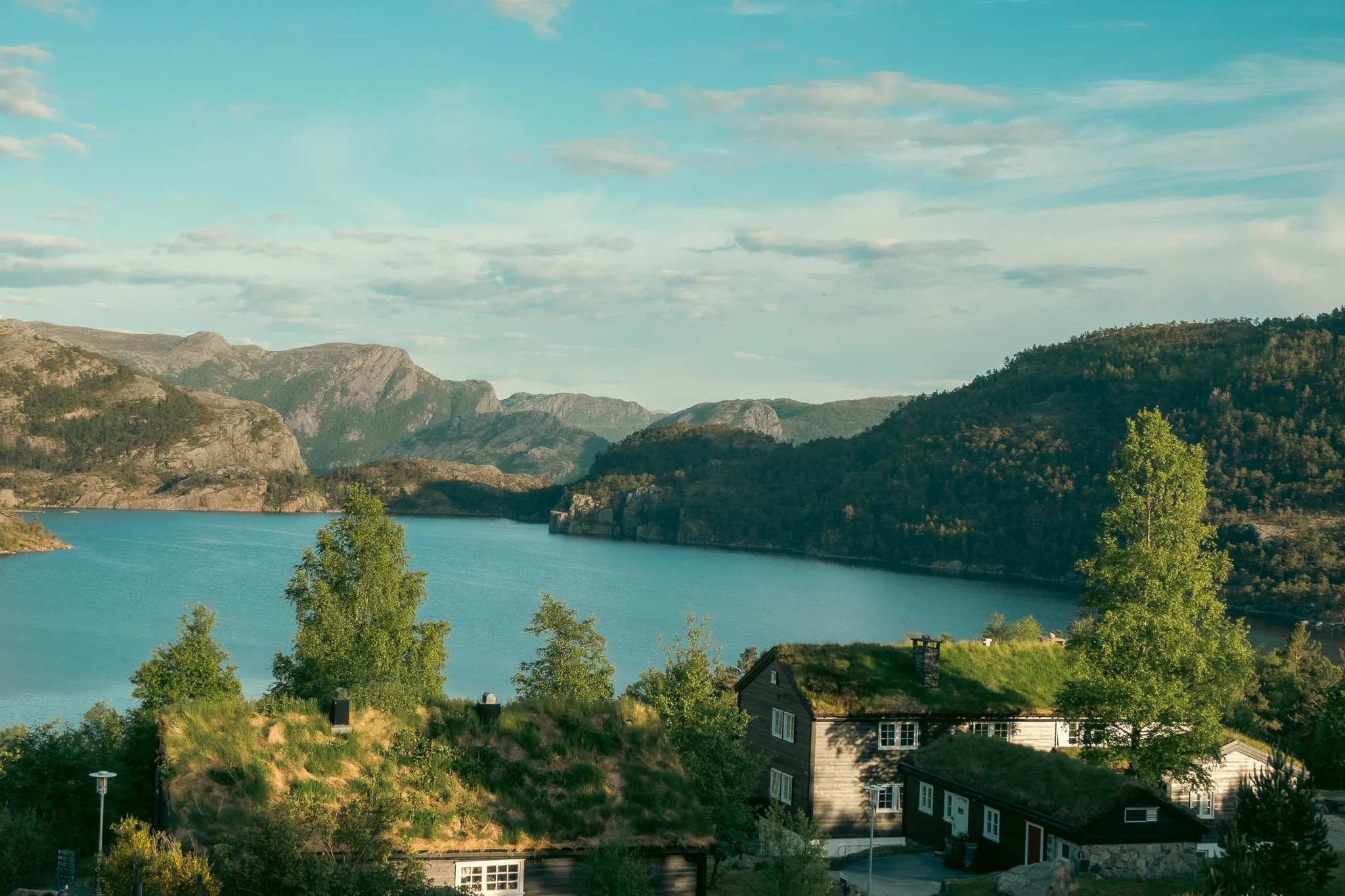 Wooden cabins lined by trees with a lake and mountains in the background on a blue sky day.