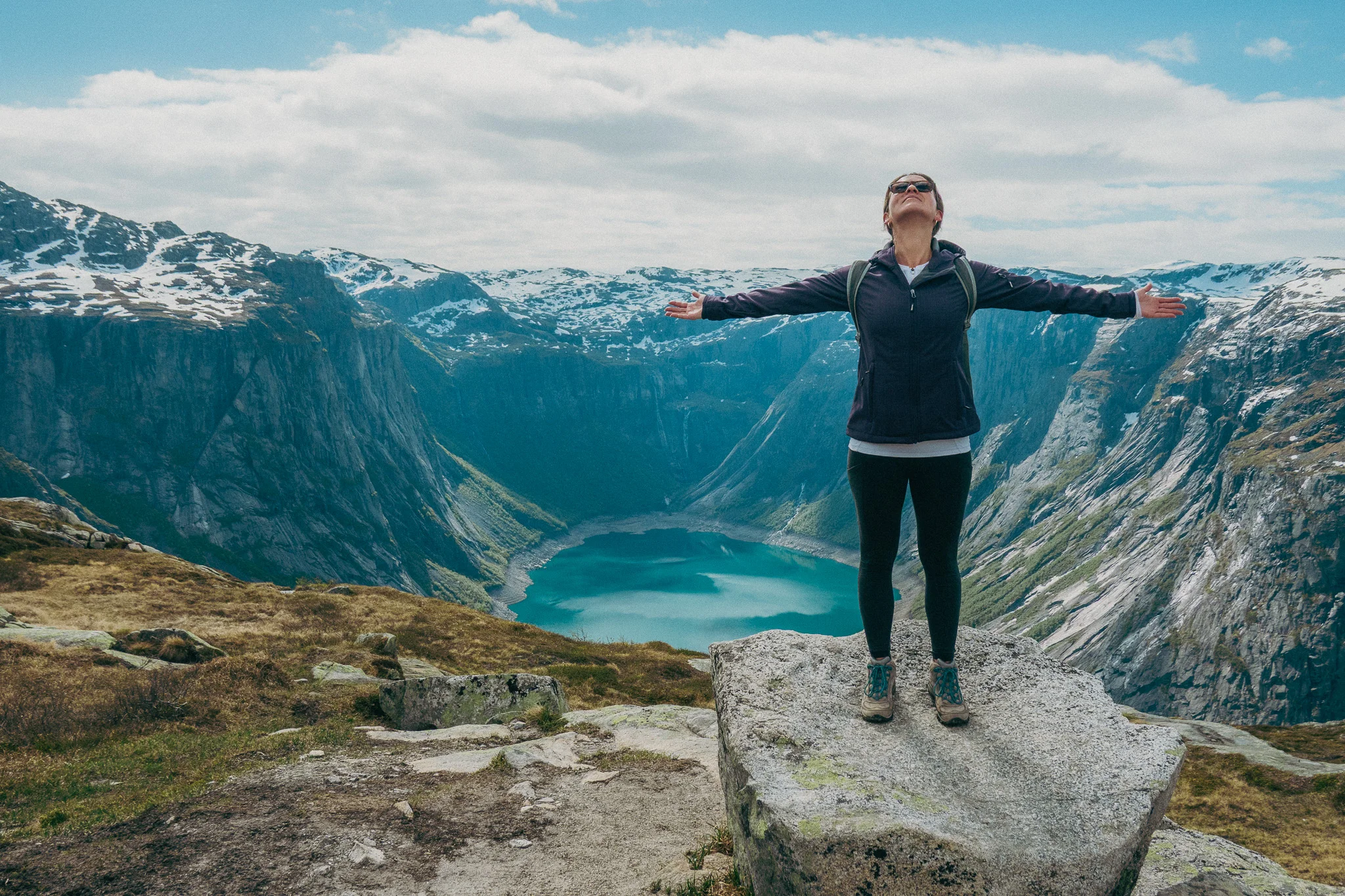 Adult female poses with arms spread in front of scenic landscape and snowcapped mountains on the Trolltunga trail in Norway