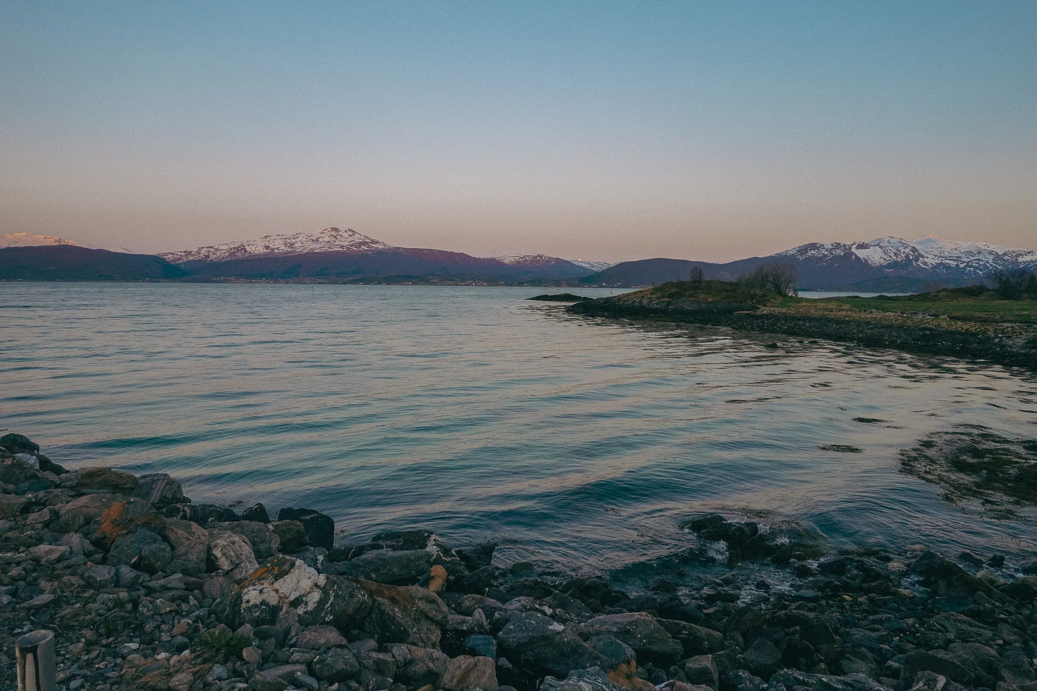 Snow-capped mountain landscape with water and rocky land in the foreground at dusk in Narvick Norway