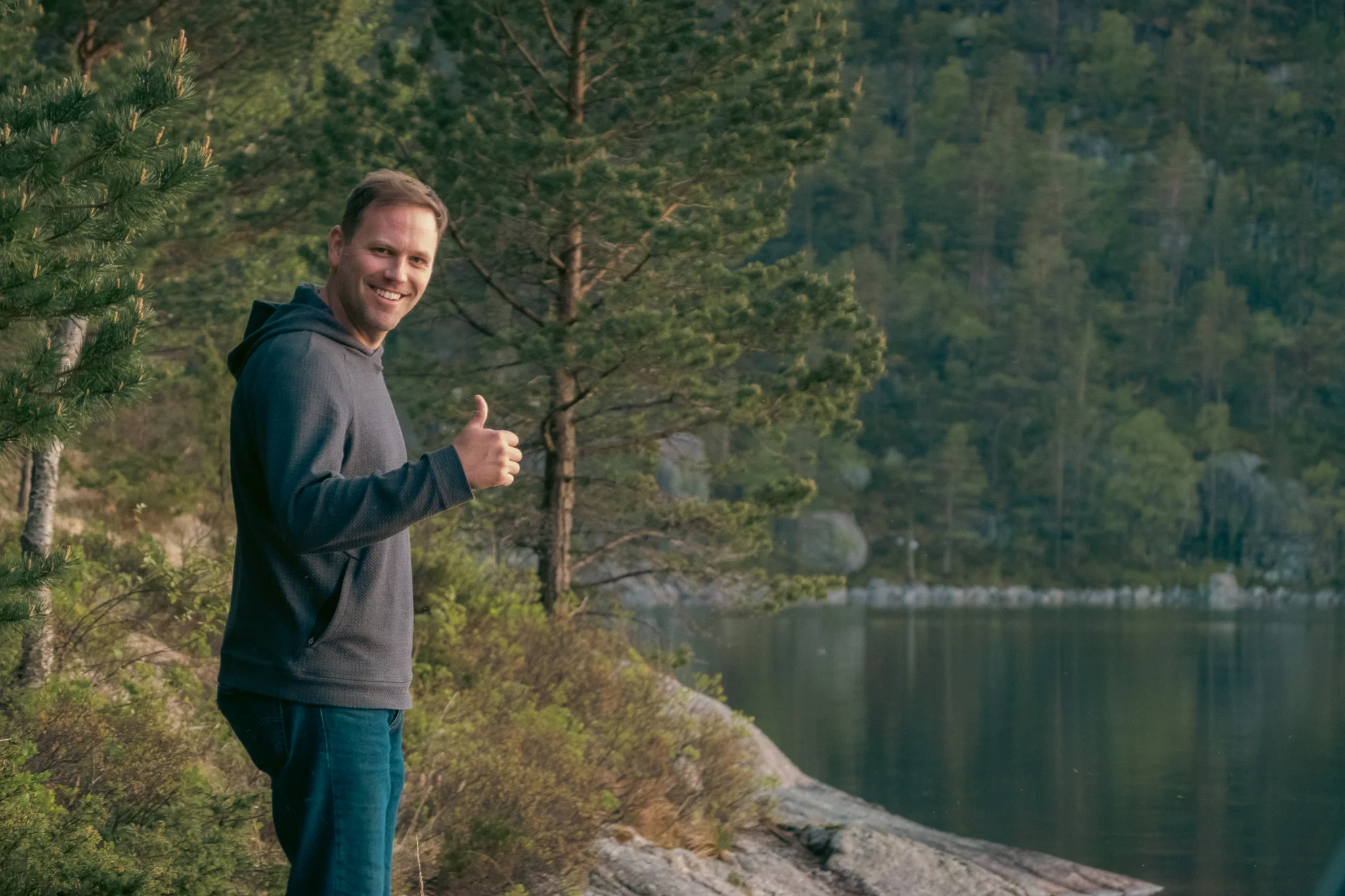 A man giving a thumbs up standing on the shore of a lake with trees and mountains in the background.