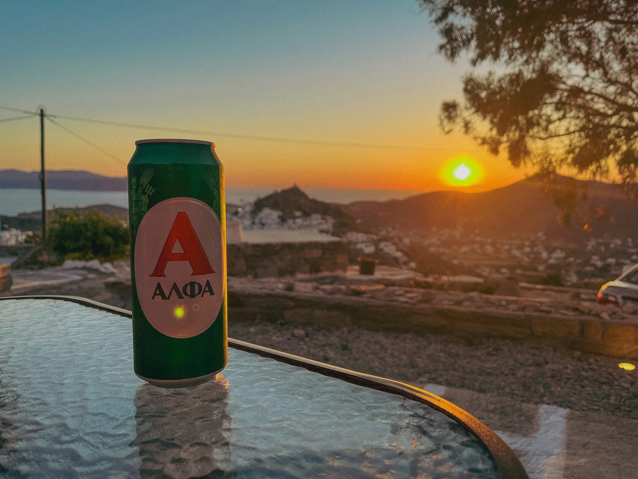 Greek Alfa beer on a glass patio table with a beautiful sunset over the mountains and water in the background