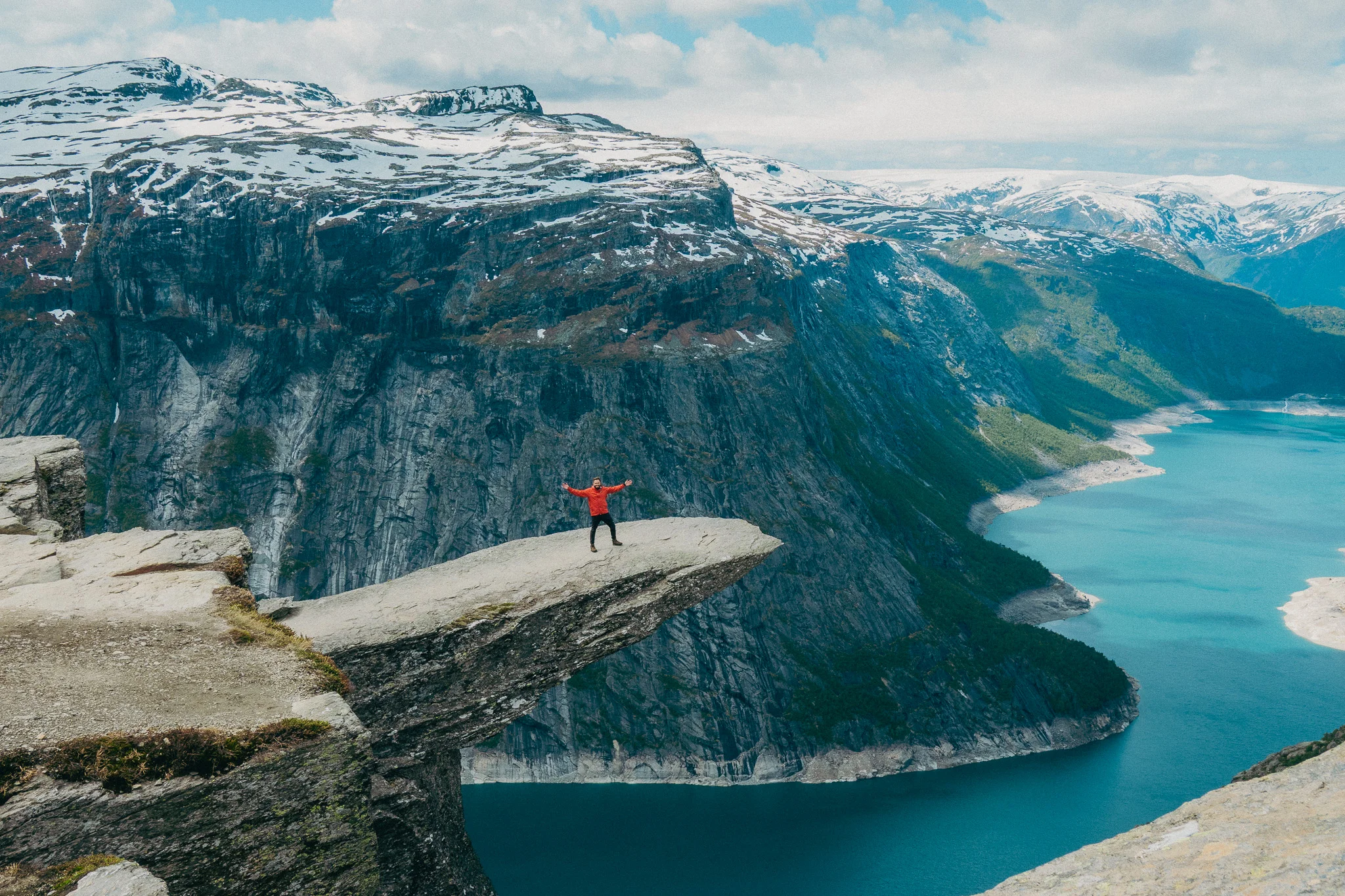 Adult male stands at the tip of Trolltunga in Norway with snowcapped mountains and blue water in the background
