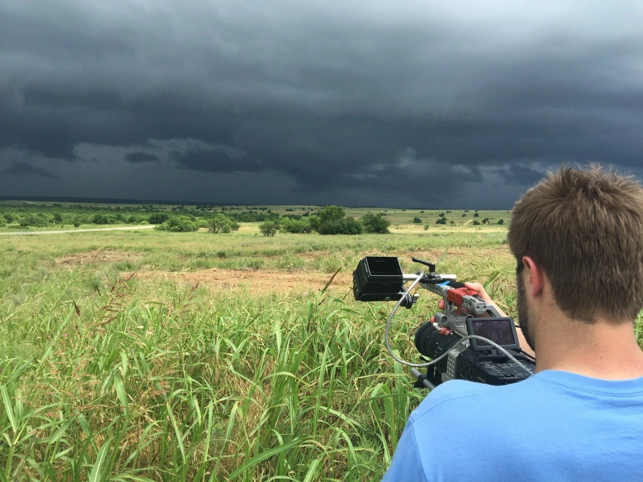 Doug Newton focusing a video camera in an open field in Oklahoma with dark stormy clouds in the background