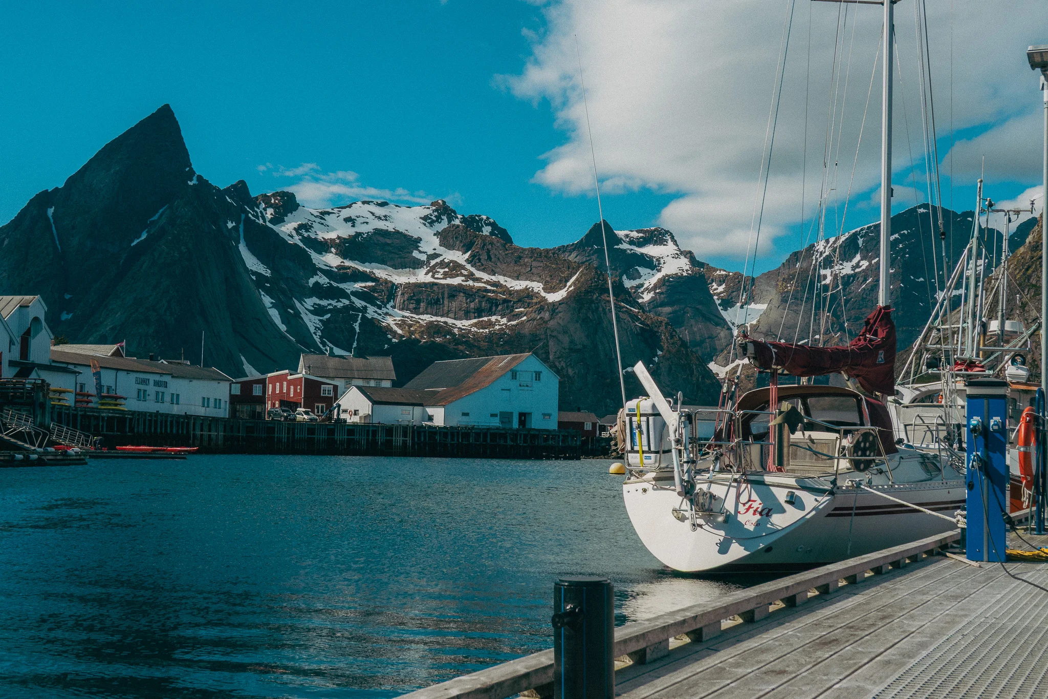 A white sailboat docked in a small marina surrounded by jagged, rocky, snow-capped mountains
