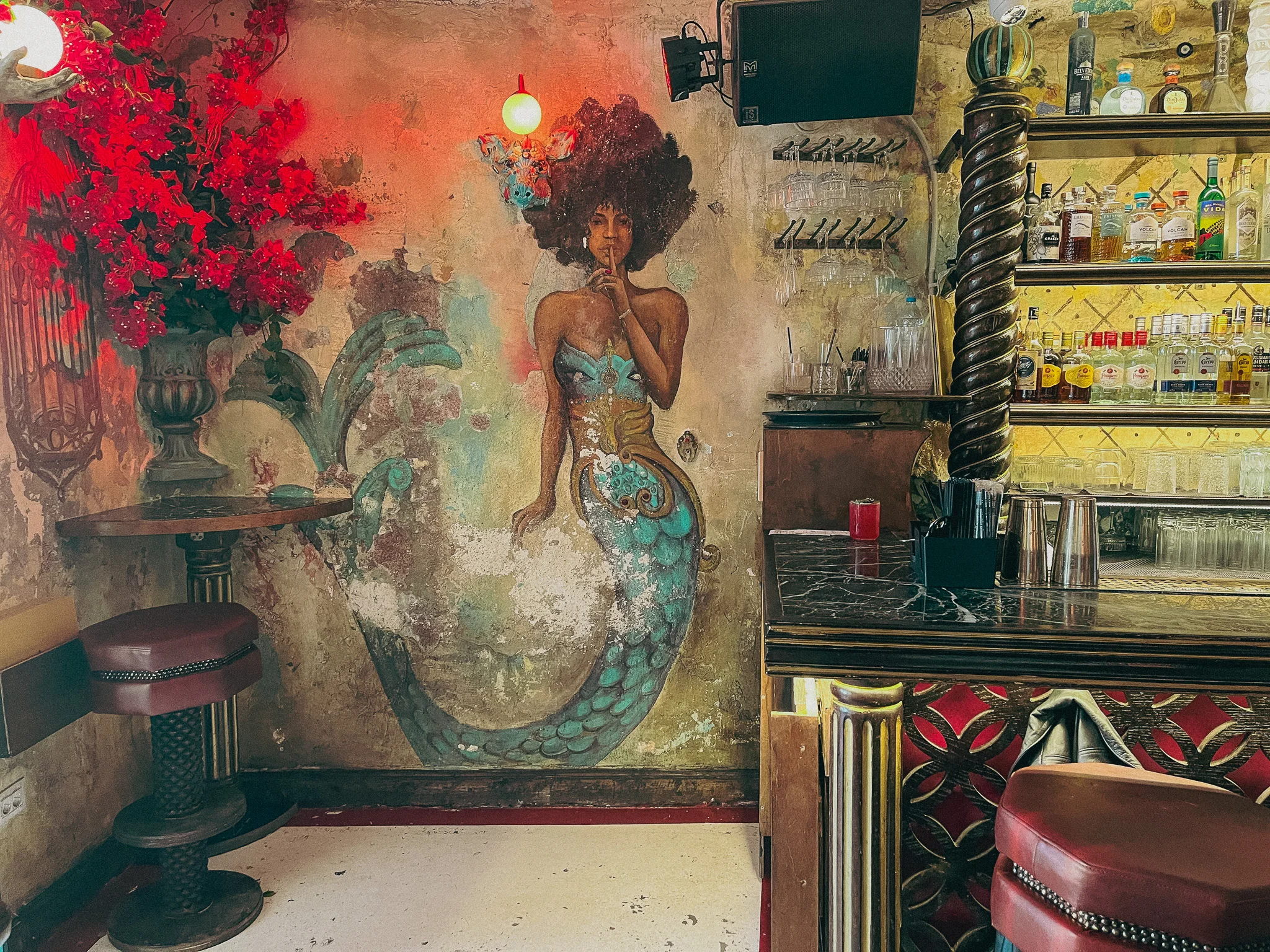 A mural on a wall of a black woman as a mermaid and black hair next to a bar and red roses as decoration in the corner