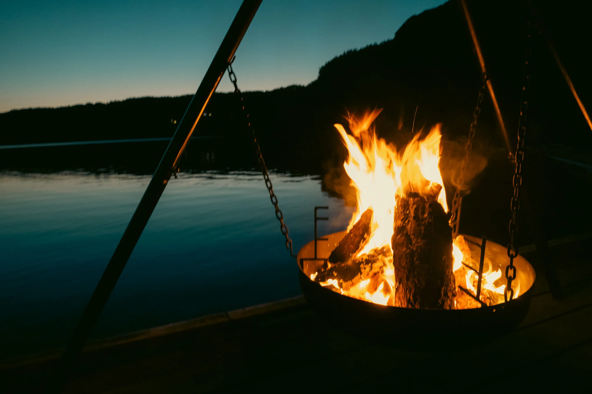 A fire burning at night alongside a lake with silhouettes of mountains in the background.