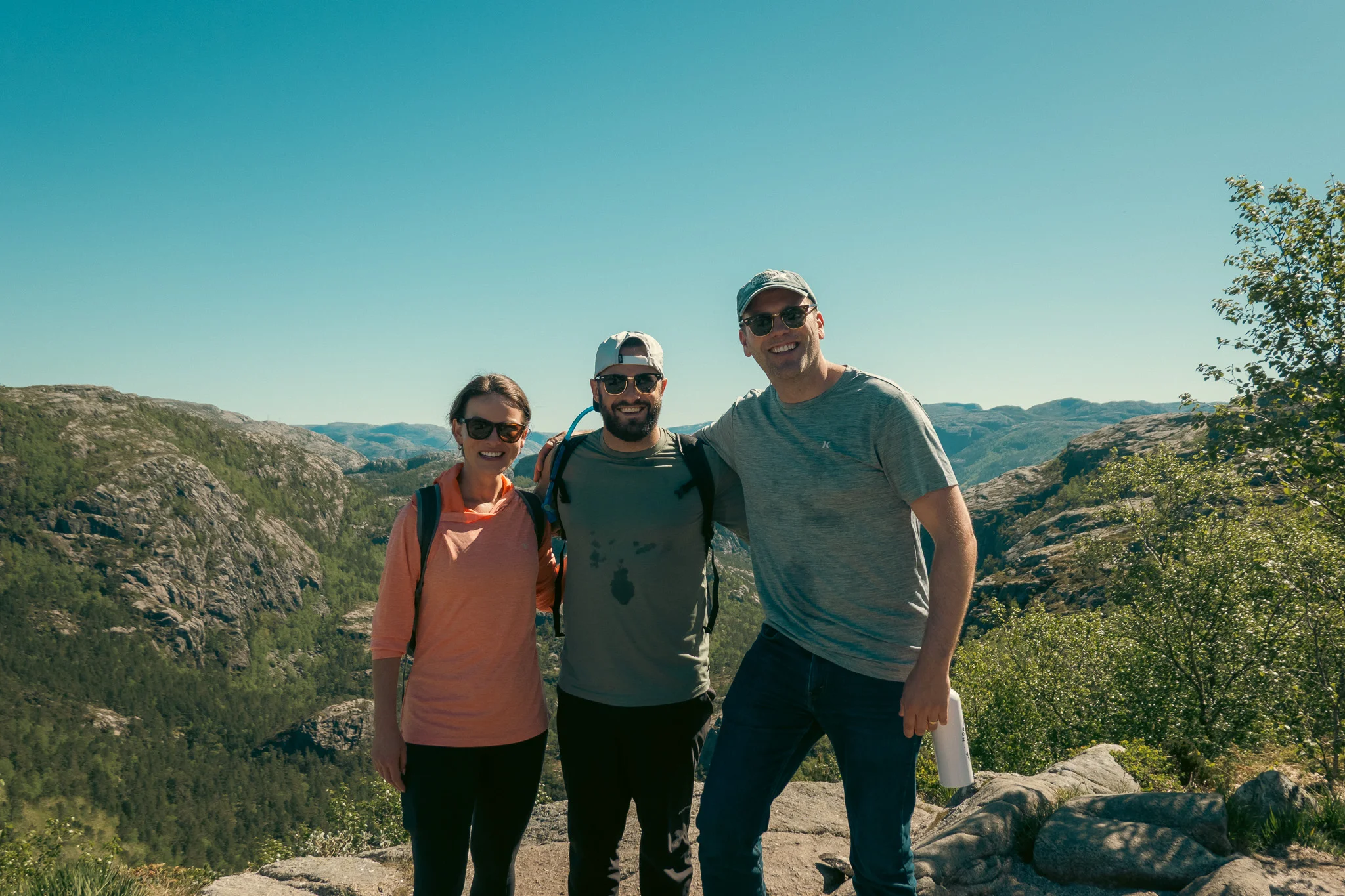 Two men and a woman pose in front of mountains and a fjord on a clear blue sky day.