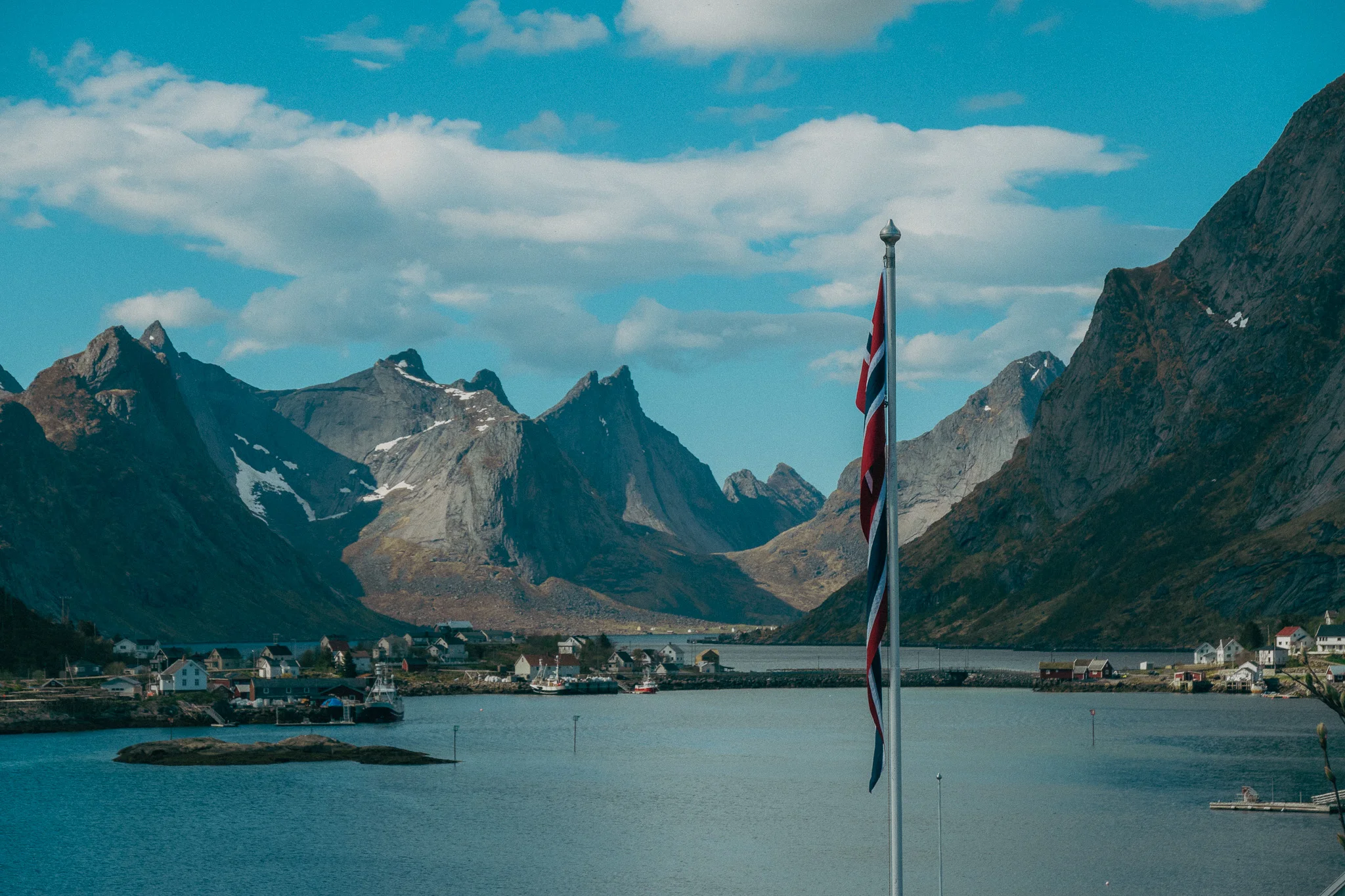 Expansive snow-capped mountains surround a small fishing village on the water with a Norwegian flag in the foreground