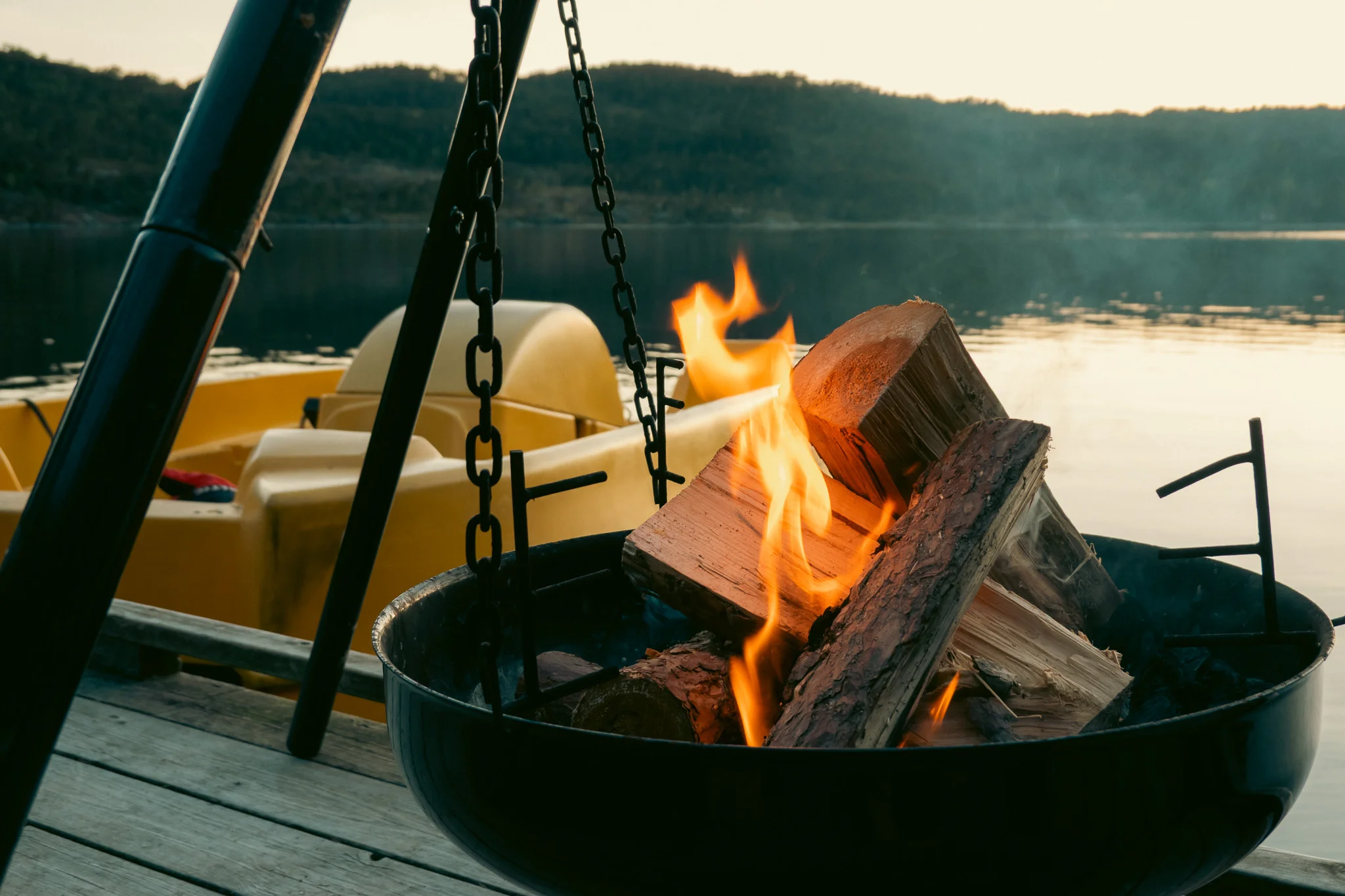 A campfire alongside a lake with a paddle boat and mountains in the background.