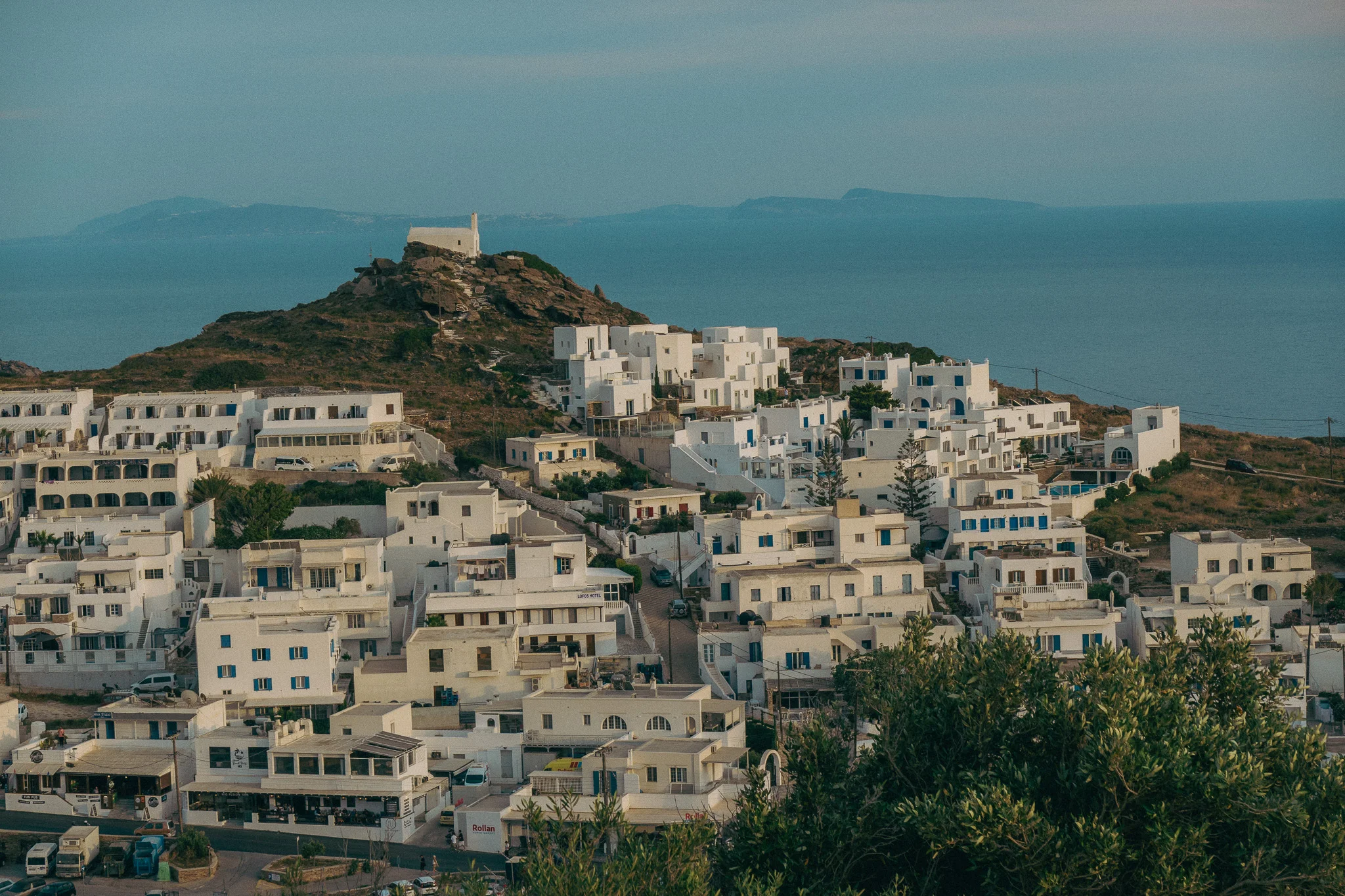 A small chapel perched atop a hill surrounded by white building and ocean in the background