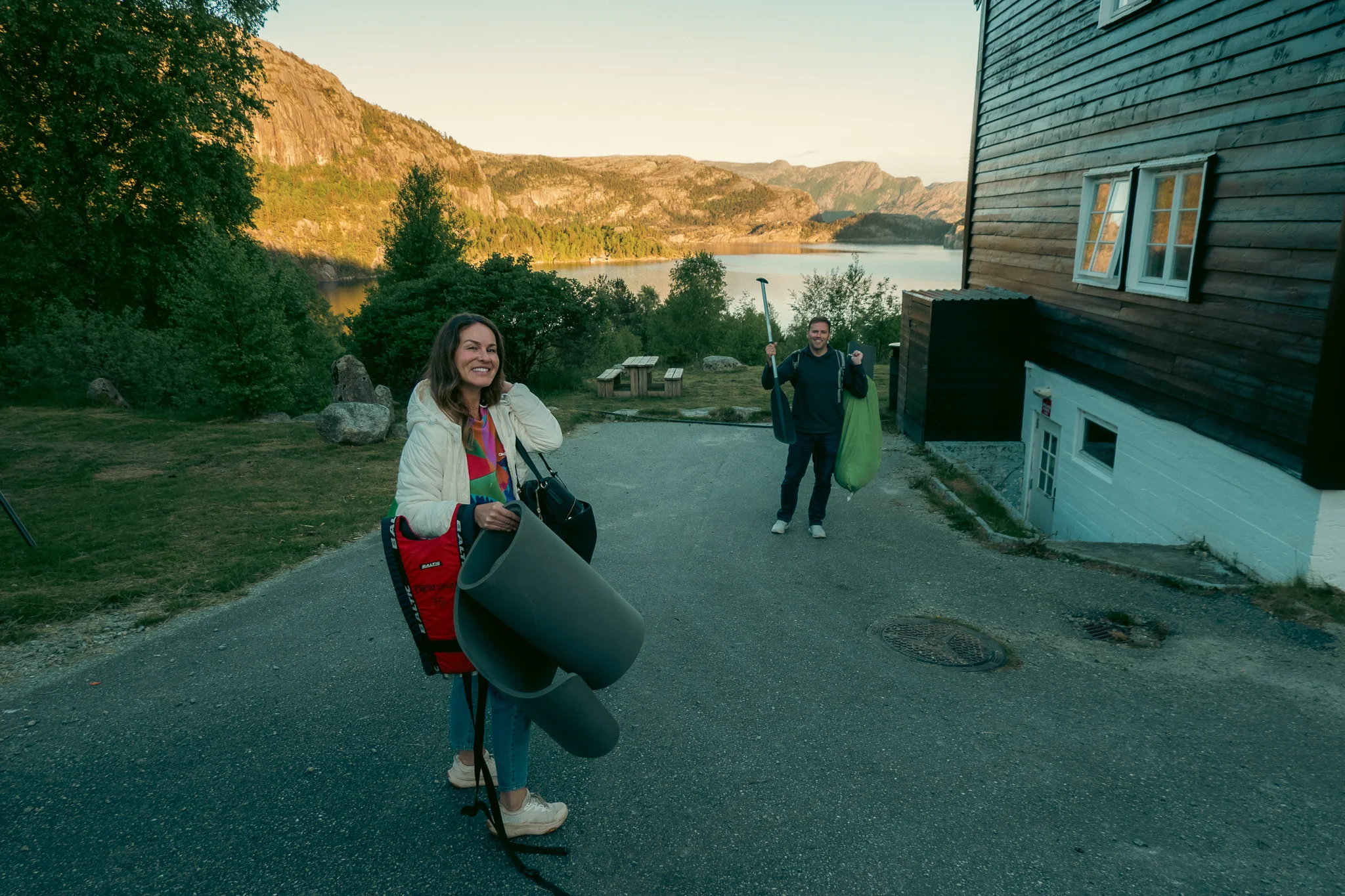 A man and woman holding camping gear and a canoe padle walking down a path towards a lake with mountains in the background at sunset.
