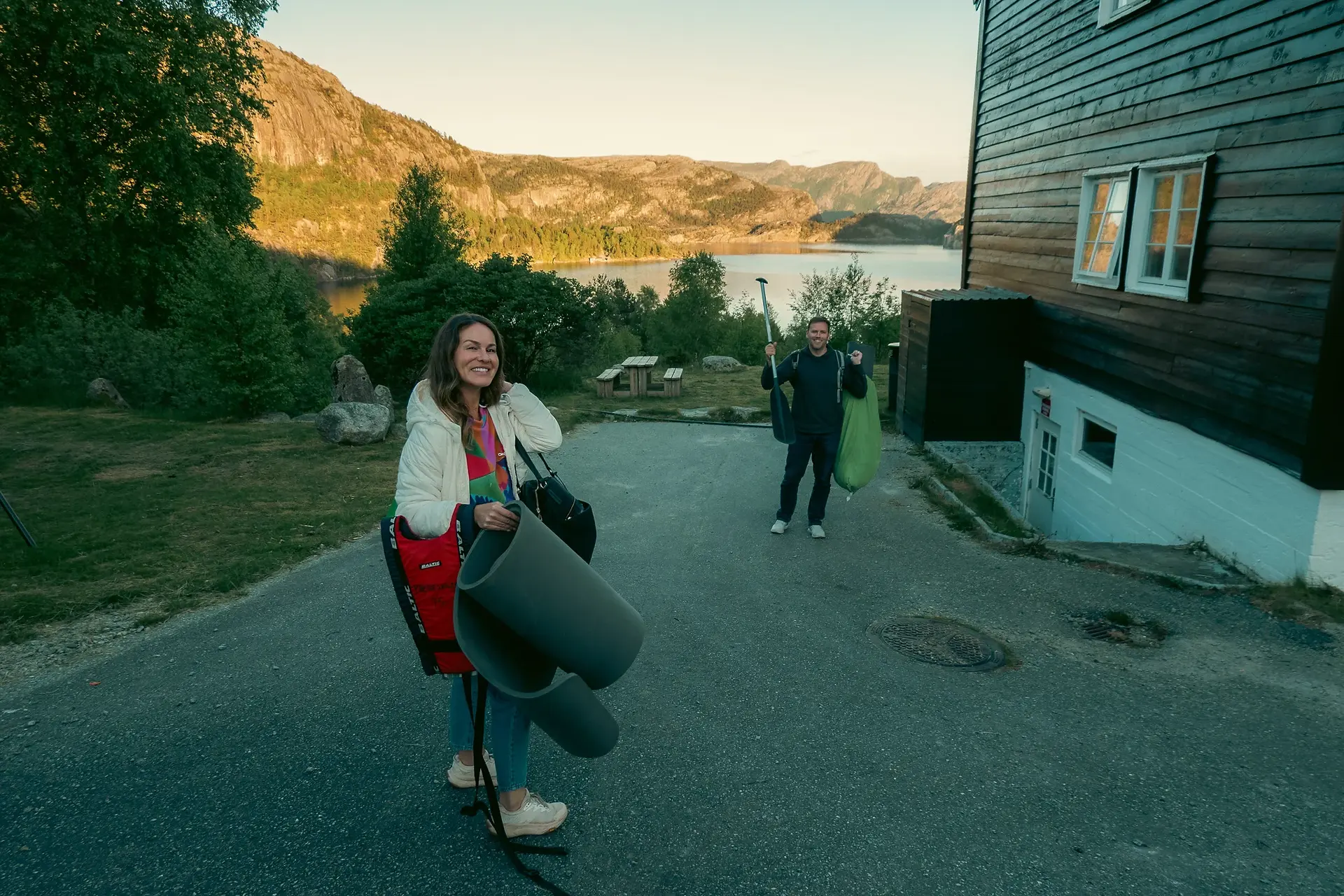 A man and woman holding camping gear and a canoe padle walking down a path towards a lake with mountains in the background at sunset.