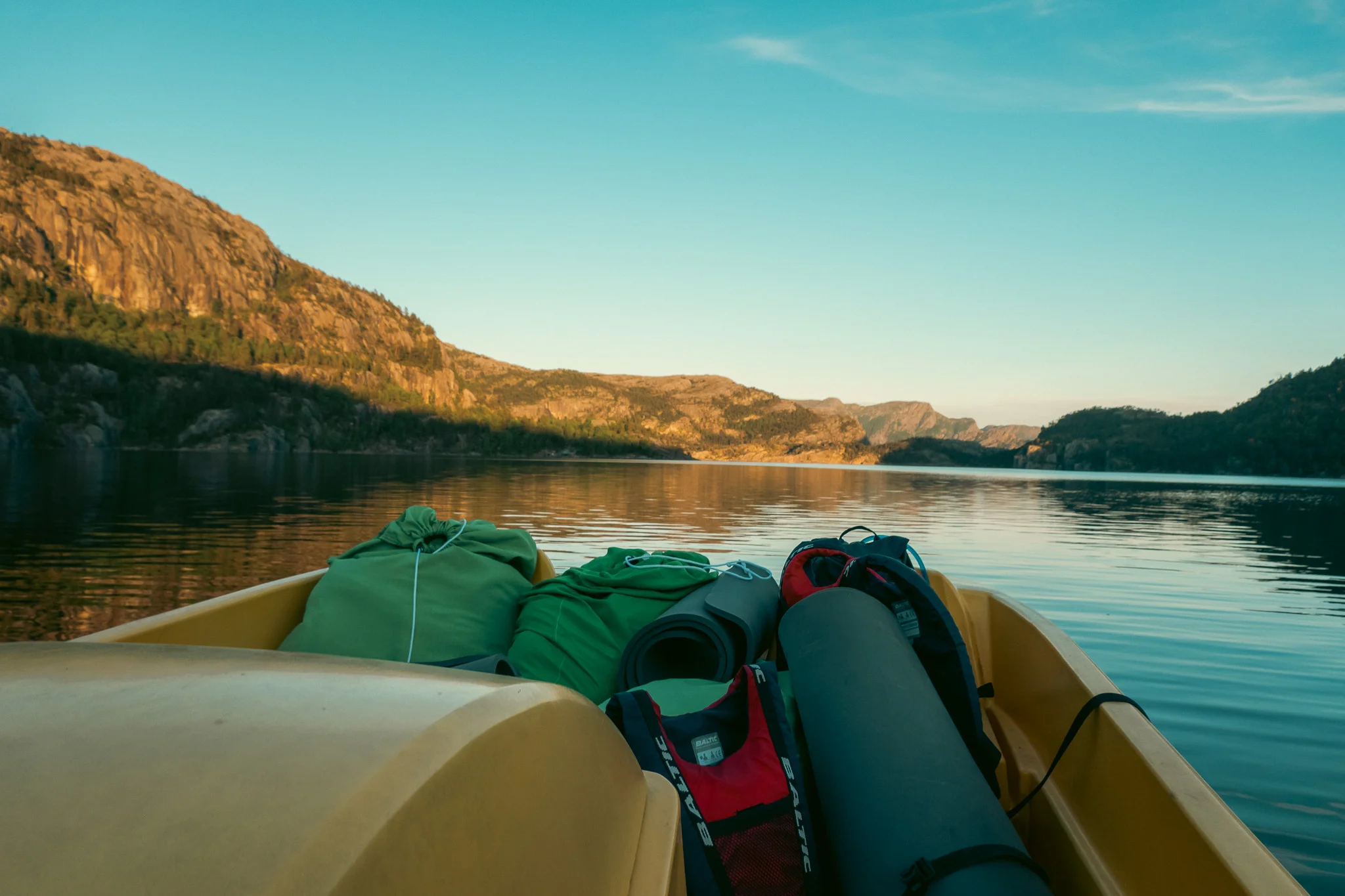Bags of gear, life vests, and sleeping pads in a paddle boat on a lake with mountains in the background at sunset.