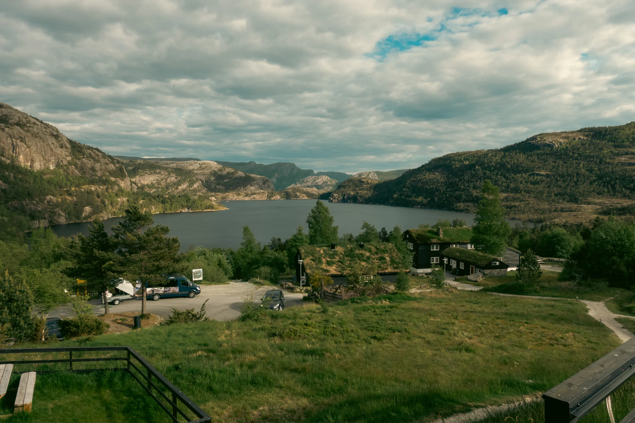 Wooden cabins lined by trees with a lake and mountains in the background on a blue sky day.