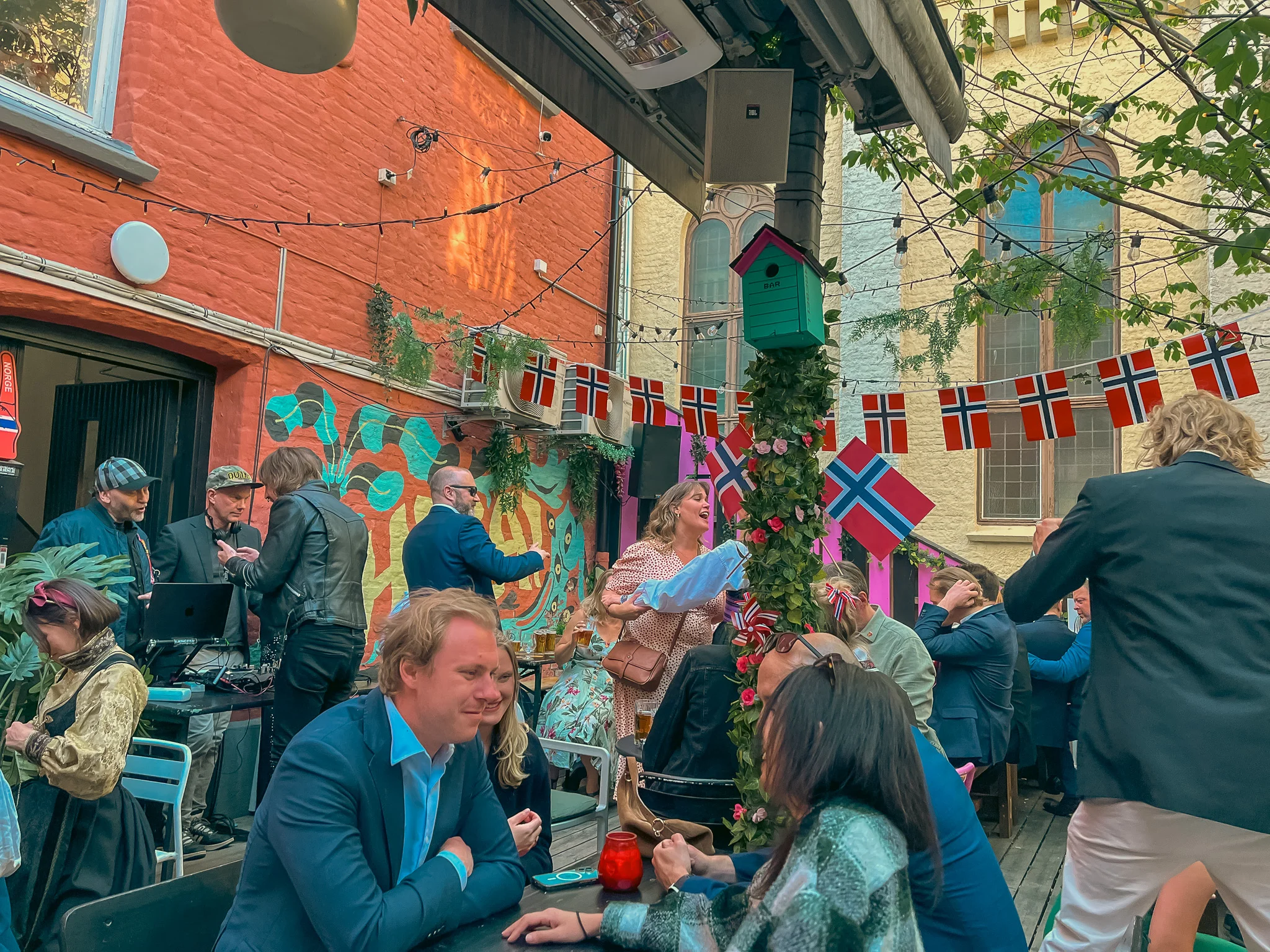 Several groups of people and the DJ on the back patio at Rebell bar in Oslo, Norway celebrating Constitution Day with Norwegian flags strung up in the background.
