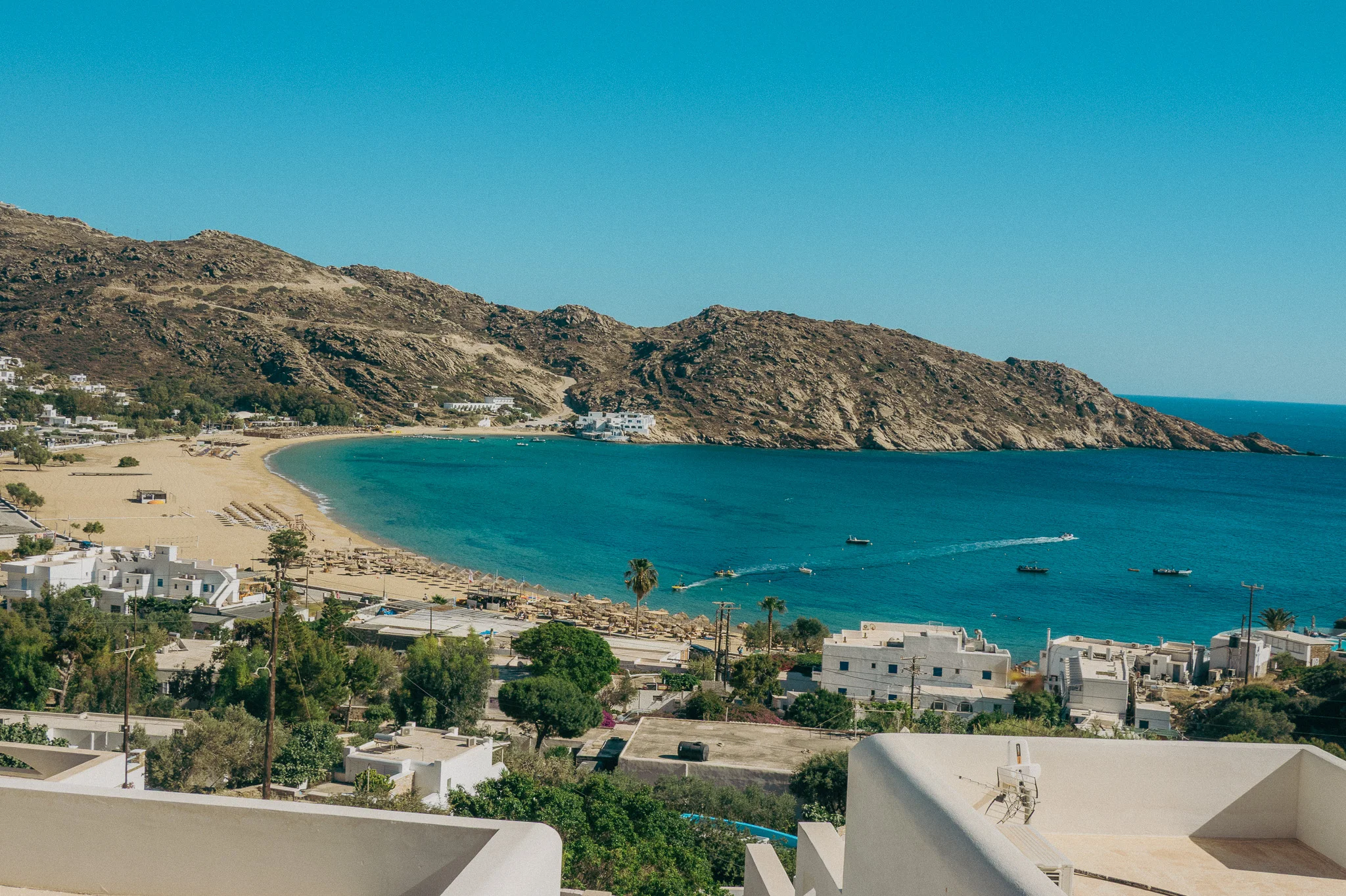 A sandy beach in Ios Greece with bright blue water surrounded by mountains, palm trees, and white buildings.