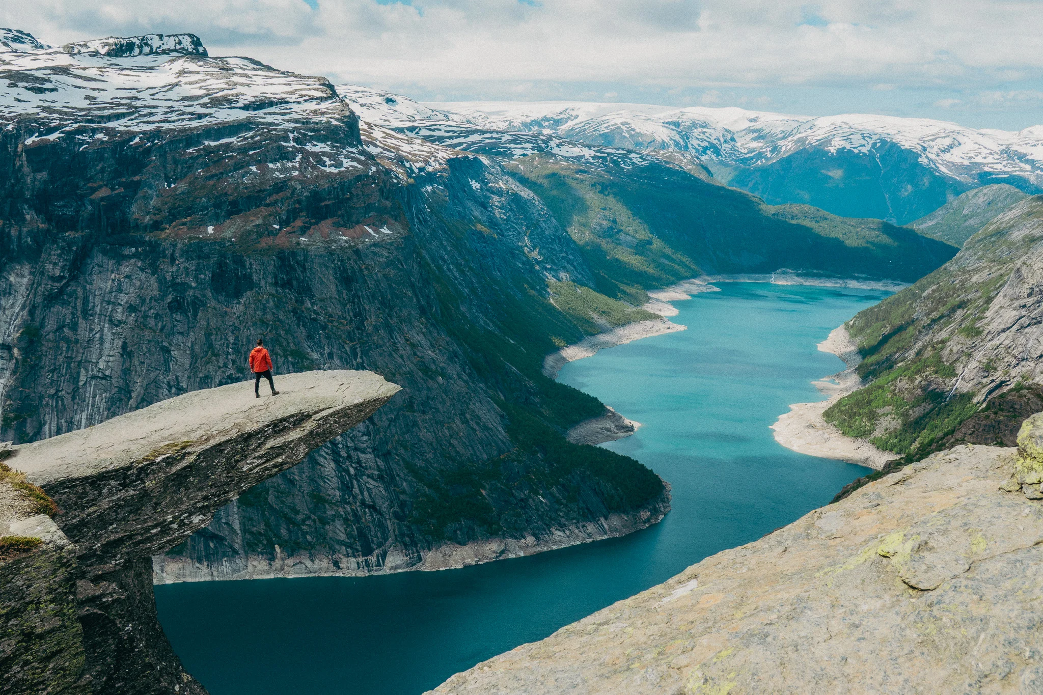 Adult male stands at the tip of Trolltunga in Norway with snowcapped mountains and blue water in the background