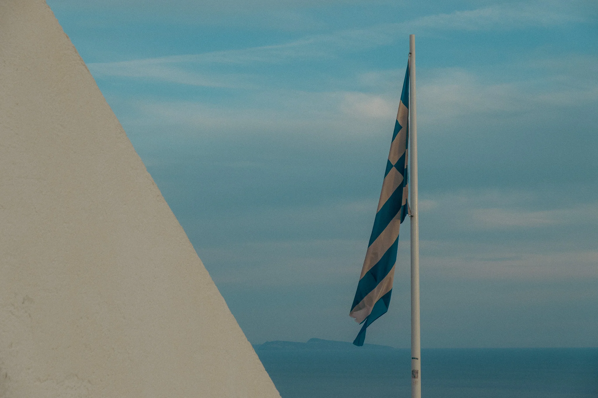 The Greek flag flying on a flag post with partly cloudy blue sky in the background at dusk