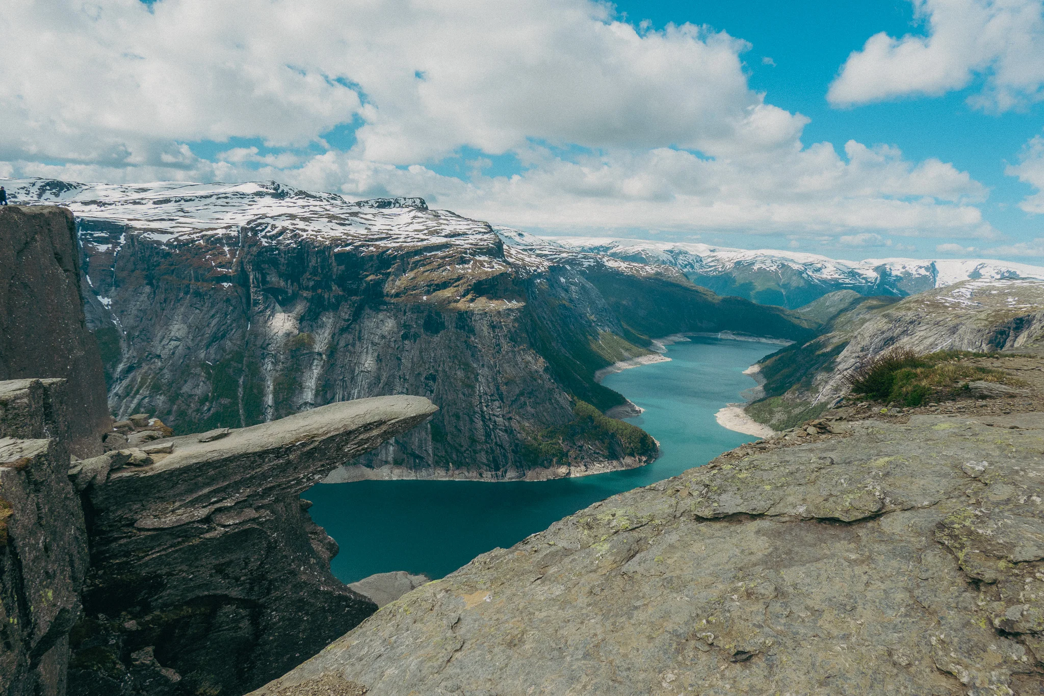 Trolltunga rock in Norway with blue water, snowcapped mountains, and partly cloudy skies in the background