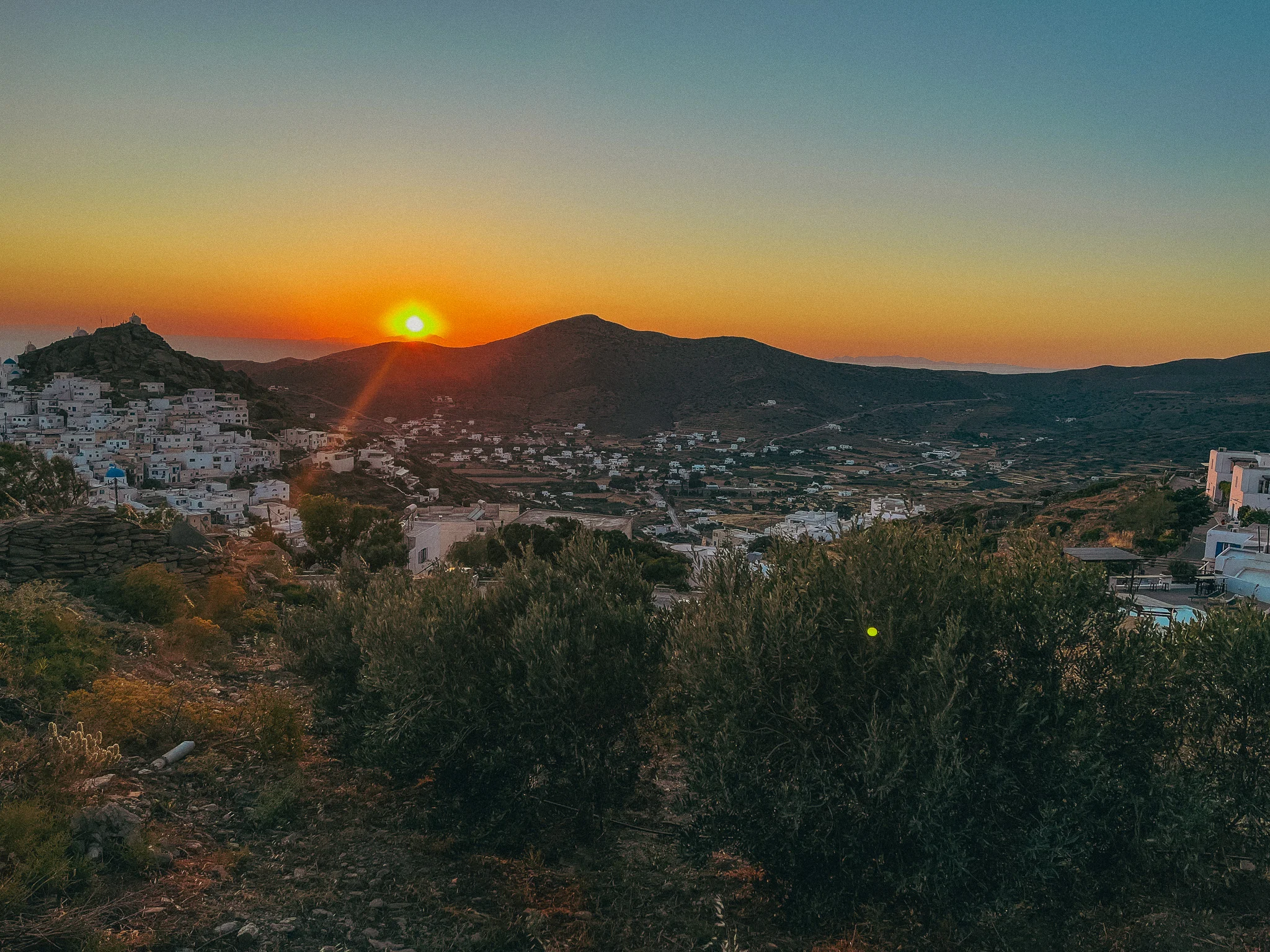 Sunset over the mountains and water with white buildings and small shrubs in the foreground